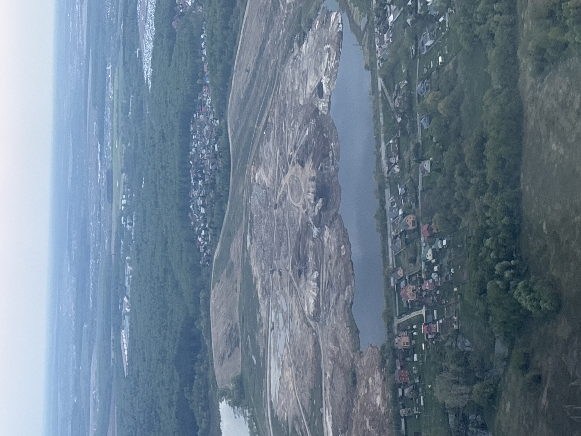 The image shows an aerial view of a landscape near the city of Domodedovo, Russia. A large quarry dominates the center of the frame, with extensive excavation and piles of material visible. A body of water runs alongside the quarry, separating it from a residential area with houses and trees on the right. In the distance, there are forests and a cityscape. The sky is overcast, and the lighting is muted. The ground appears to be dry and dusty. The image doesn't show any people or interactions.