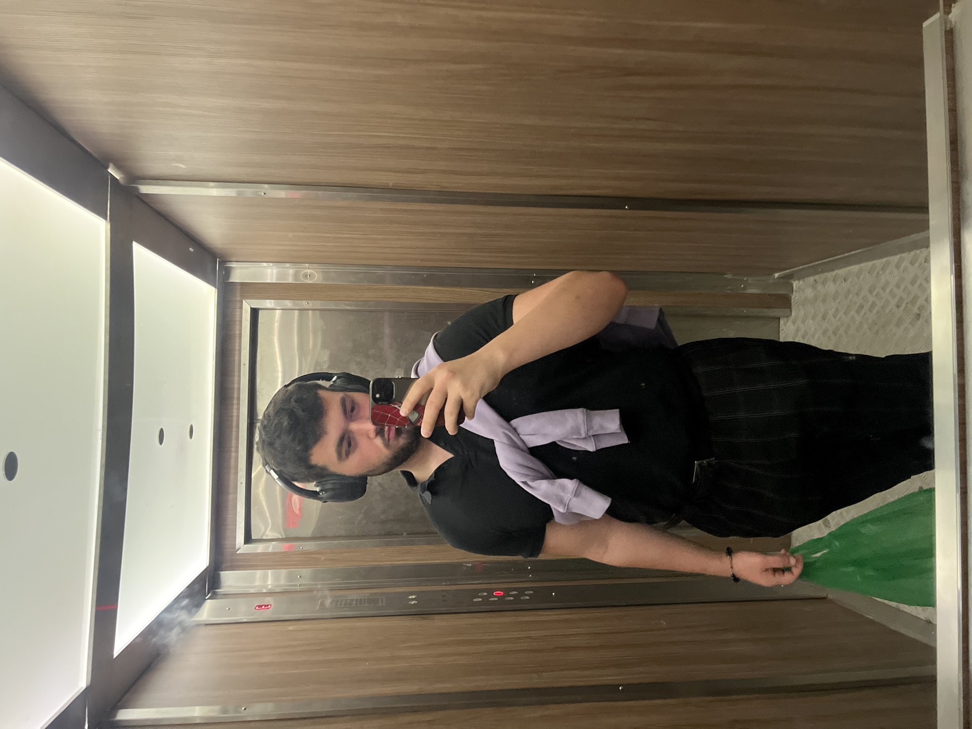 A man takes a selfie in an elevator with wood paneling and a stainless steel accent wall. He wears a black t-shirt, dark pants, headphones, and a light purple sweater is draped over his shoulders. He is holding a cell phone in his right hand. A green item is visible in the mirror reflection. The location cannot be determined as Sofia, Bulgaria, from the given image.