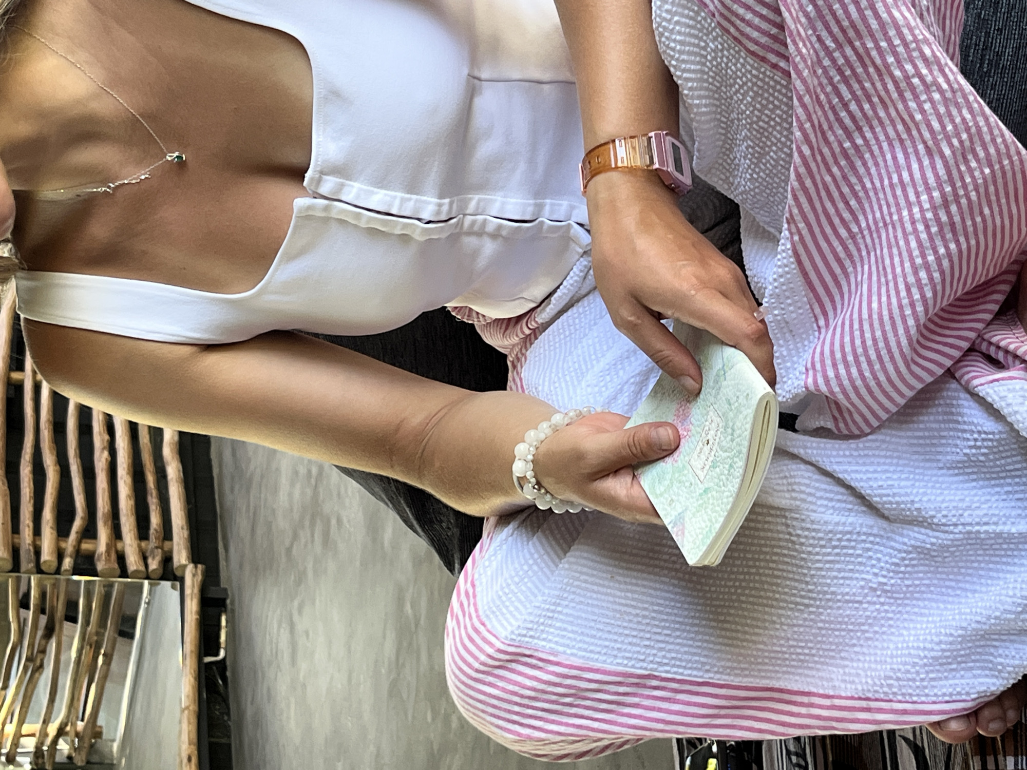 The image shows a close-up of a person from the chest down, holding a small book or pamphlet. They are wearing a white top with a neckline and a strap visible. A delicate silver necklace with a small green stone pendant hangs around their neck. On their left wrist, they wear a rose-gold and translucent pink watch and a bracelet made of small white beads. The person is partially wrapped in a white and pink striped fabric, possibly a sarong or towel, which has a textured seersucker pattern. The background is out of focus, but appears to be a textured wall. The lighting suggests it might be daytime. The person is holding the book open with their right hand, showing pages with a floral print and some faint text. The overall impression is a candid shot of someone in a casual setting, possibly a beach or resort, given the attire and the striped fabric.