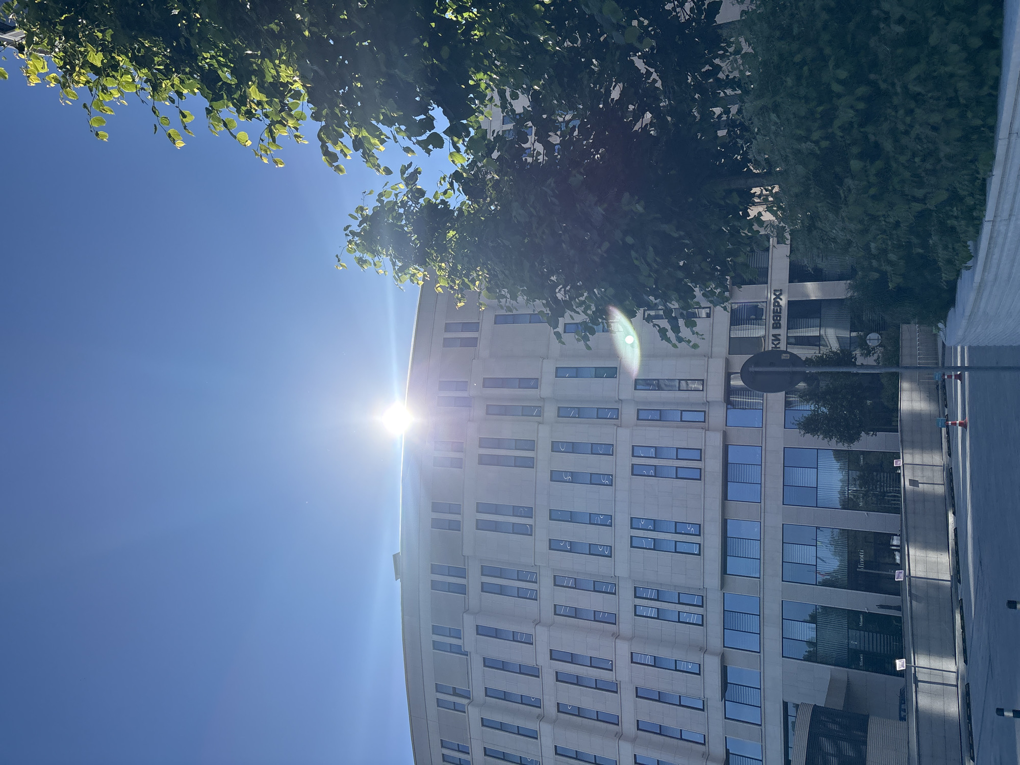 This is a photograph of a curved, modern office building in Minsk, Belarus, taken from a low angle on a sunny day. The sun is positioned directly above the building, creating a bright lens flare that obscures the top portion of the structure. The building's facade is composed of light-colored stone and features numerous rectangular windows, many of which appear to have blinds or shutters. On the ground floor, the building has a glass entrance with signage visible. Green trees and bushes are in the foreground and partially obscure the building. The sky is a clear, bright blue. The image captures a portion of a street or sidewalk in front of the building, with concrete barriers and a traffic cone visible. The overall impression is one of a well-maintained, contemporary urban environment.