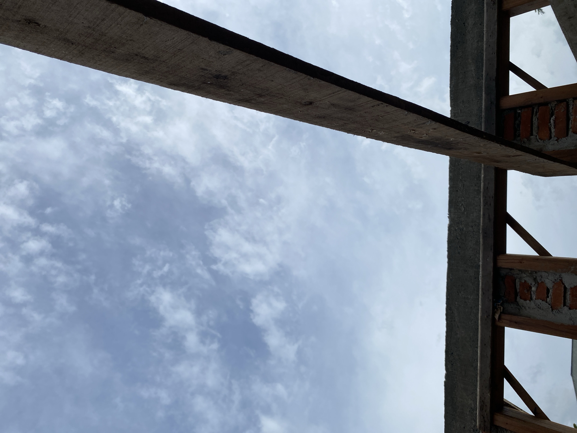 The image shows a construction site with a cloudy sky. In the foreground, there is a wooden beam crossing diagonally across the frame. Below the beam, there is a concrete structure with exposed brickwork and wooden framing. The sky is filled with scattered clouds, appearing light blue and white. The scene suggests an unfinished building or renovation project. The location is described as Unknown, Unknown.