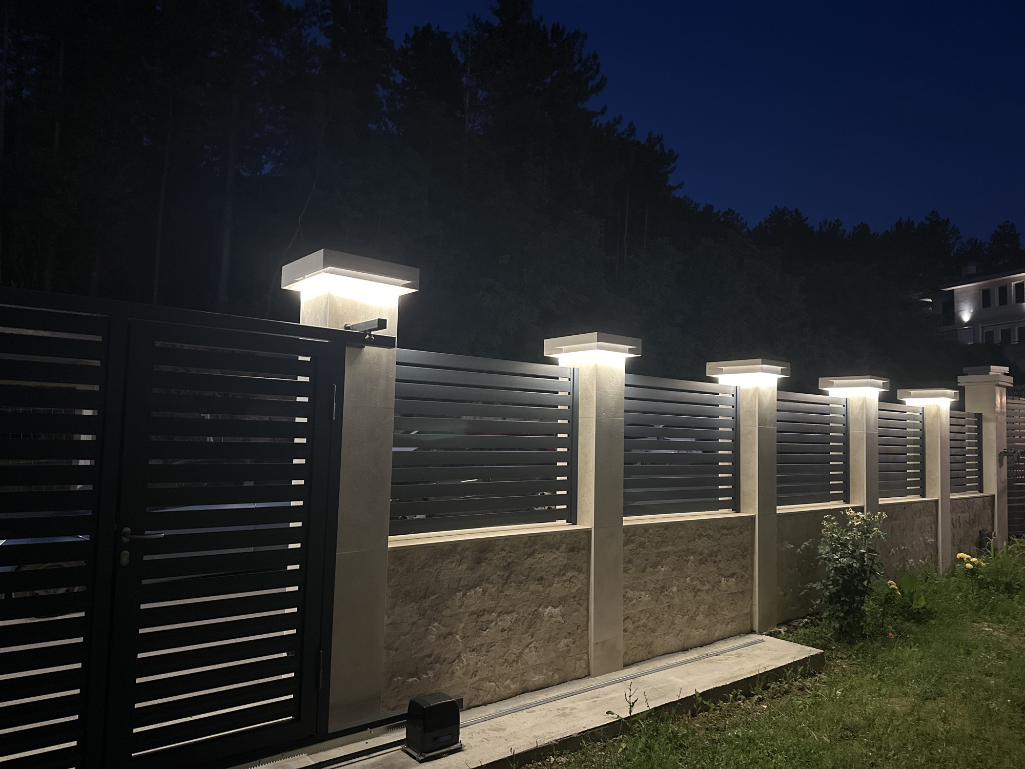 The image displays a modern perimeter fence and gate illuminated at night, located in Asenovgrad, Bulgaria. The fence consists of dark-colored horizontal slat panels, interspersed with and supported by light-colored stone or concrete pillars. Each pillar is topped with a rectangular light fixture emitting a warm, upward glow. Below the slat panels, the fence features a lower wall section constructed from textured, light brown stone blocks.
On the left, a large sliding gate, matching the horizontal slat design, is visible. A smaller pedestrian gate is integrated into this larger structure, featuring a visible handle. At the base of the sliding gate, a black automated gate operator motor is positioned on a concrete track.
The background reveals a dense, dark forest covering a hillside under a deep blue night sky. To the far right, partially obscured by trees, a multi-story building with illuminated windows and exterior lights is visible in the distance.
In the foreground, a strip of unkempt grass and some sparse vegetation, including a small bush and several yellow-flowering plants, runs along the base of the fence. A concrete edge or path is present at the immediate base of the fence structure. No individuals are present in the scene.