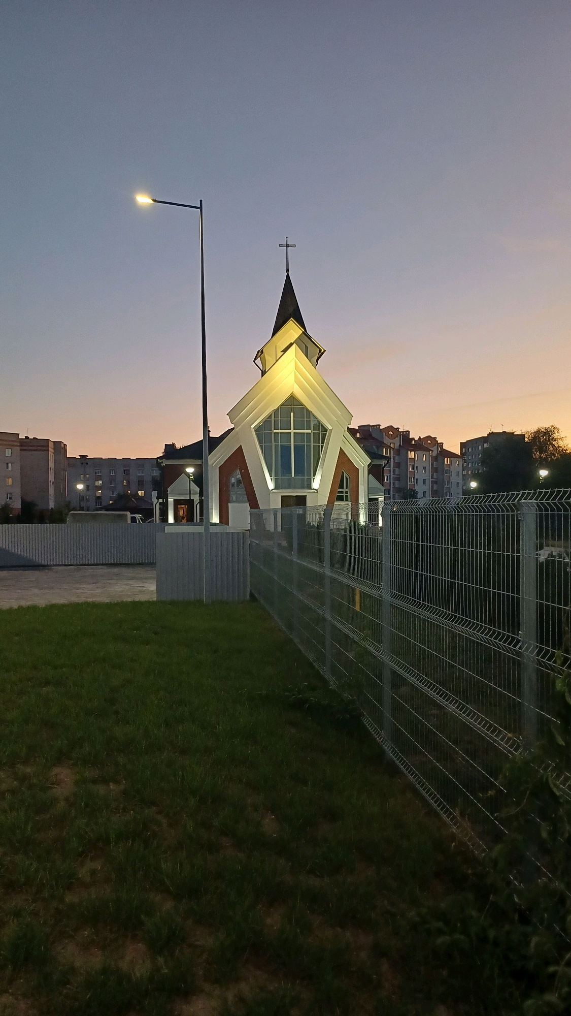 The image depicts the exterior of a modern church building at dusk. The church has a distinctive design with a white, angular facade and a dark steeple topped with a cross. The building is illuminated by external lights, highlighting its architectural features. In the foreground, there is a well-maintained lawn and a metal fence. Beyond the church, several apartment buildings are visible, characteristic of a residential area within a city. A street lamp stands to the left of the church, casting a pool of light on the ground. The sky is a gradient of twilight colors, transitioning from pale blue to soft orange. This scene is located on Ryabceva Street in Brest, Belarus. There are no people or animals visible in the image.