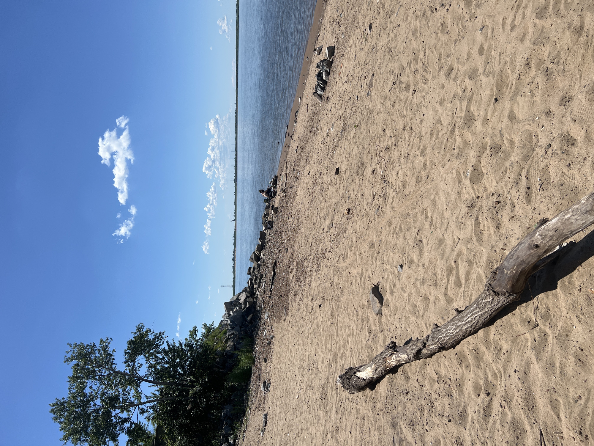 The image shows a sandy beach with a large piece of driftwood in the foreground. The beach is next to a wide body of water, likely a river, with a faint line of trees and industrial structures visible on the opposite shore. The sky is blue with scattered white clouds. In the mid-ground, a person is sitting on the rocky edge of the beach, facing the water. The scene appears to be a natural or semi-natural area near an urban or industrial zone.