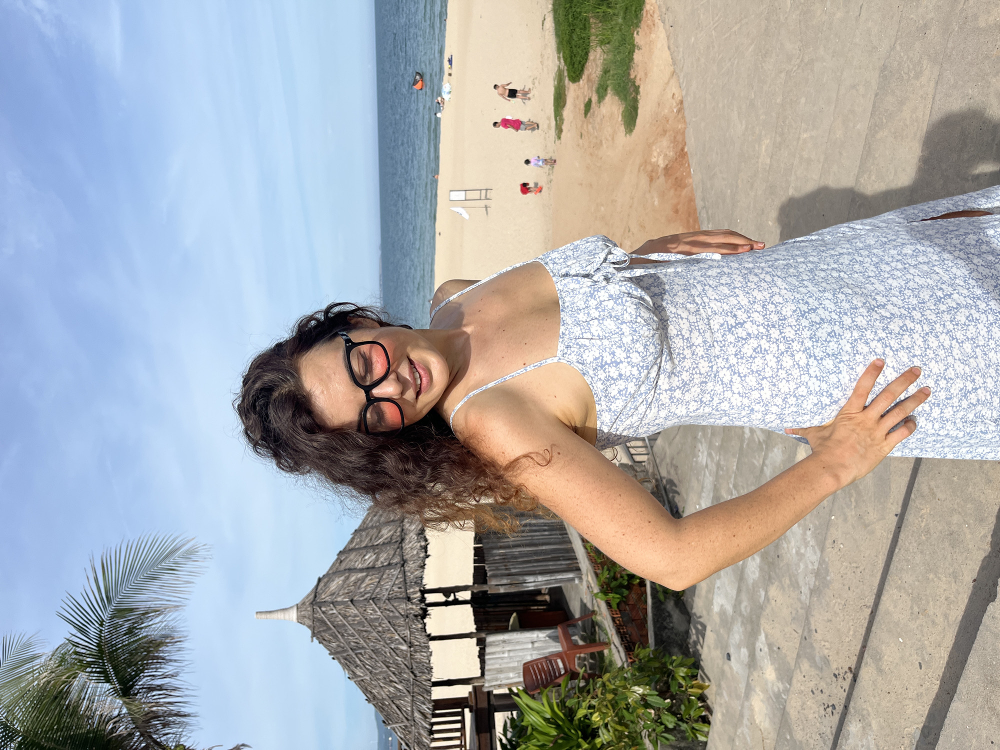 A woman with curly brown hair and dark-rimmed glasses with pink lenses stands on concrete stairs overlooking a beach in Mũi Né, Vietnam. She is wearing a light blue and white floral print dress with spaghetti straps and a tie detail at the bust. Her right hand is on her hip, and her left hand is resting on her thigh. In the background, a thatched-roof hut is visible, along with palm trees, the sea, and a sandy beach where several people, including children, are present. The sky is blue with some scattered clouds.