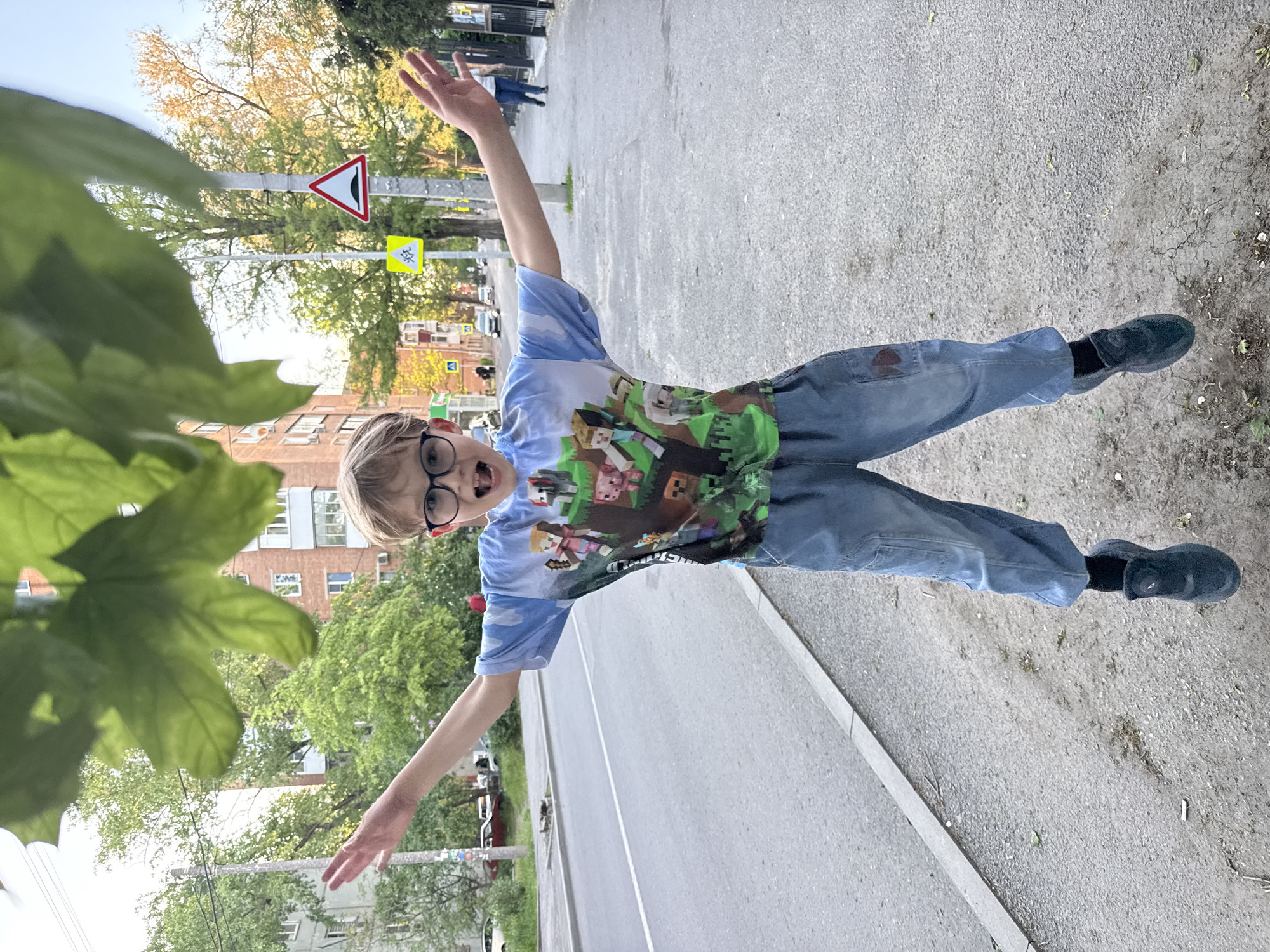 A full-body shot shows a young boy with blond hair, wearing glasses, a Minecraft t-shirt, and blue jeans, striking a playful pose with outstretched arms. He is standing on a sidewalk next to a road. A traffic sign warning of a junction is visible in the background. The location is a street in Rostov-on-Don, Russia. Buildings and trees line the street. Green foliage is partially obscuring the view from the left side. The pavement is gray.