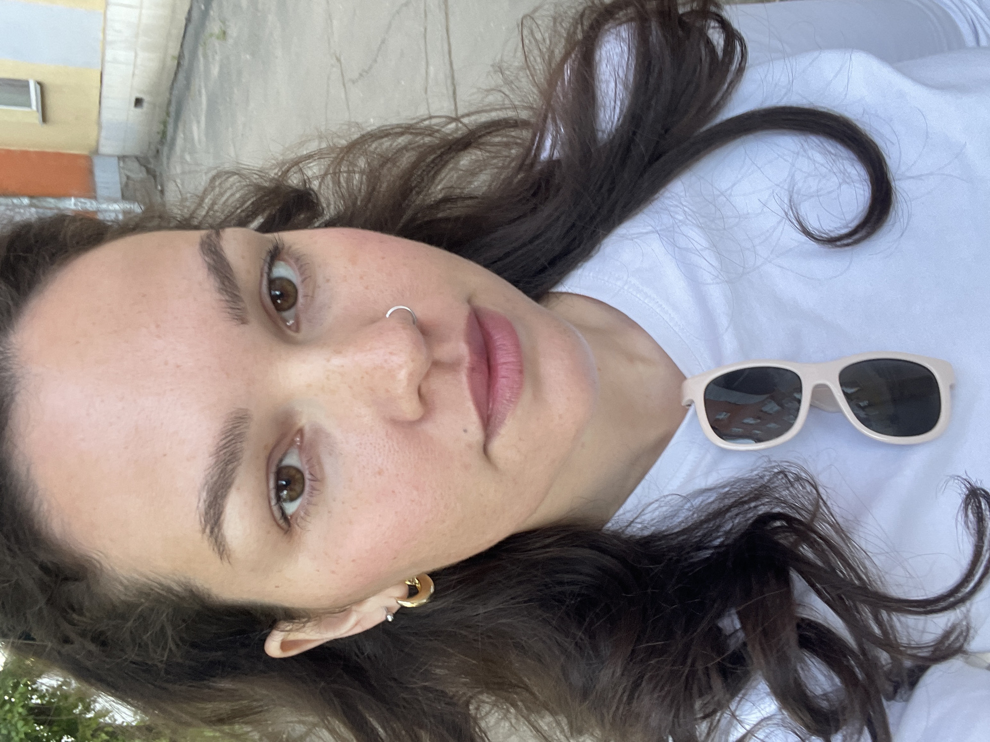 A close-up, tilted portrait shows a young woman from the chest up, likely taking a selfie outdoors in Izhevsk, Russia. She has fair skin with visible freckles, dark brown eyes, and full, dark eyebrows. A small silver hoop nose ring is in her left nostril, and her left ear is adorned with a prominent gold hoop earring and a smaller silver stud below it. Her lips are a natural pink hue. Her dark brown, wavy hair is spread around her shoulders and across the white t-shirt she is wearing. Resting on her shirt to her right are a pair of light-colored (possibly beige or off-white) sunglasses with dark lenses.
The background is softly blurred but reveals an urban outdoor environment, consistent with Izhevsk, Russia. On the left, parts of a building with yellow and orange sections and a small window are visible. Behind her head and hair, there appears to be a concrete or paved surface. The bright, natural lighting indicates it is daytime, and the presence of sunglasses suggests sunny weather. The woman maintains a calm, neutral expression, looking directly towards the camera. No visible text is present in the image.