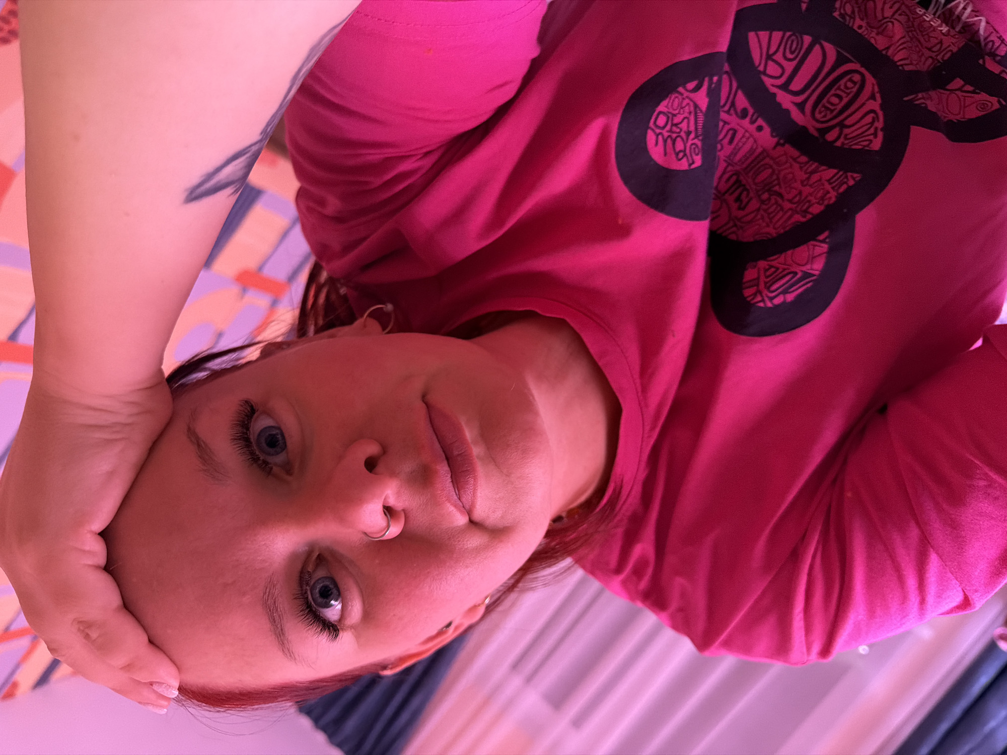 The image captures a close-up, low-angle perspective of a female subject situated indoors in the city of Chaikovsky, Russia. The subject, possessing reddish hair and blue eyes, directly gazes at the camera. Prominent false eyelashes are visible, along with a silver septum ring. Her right hand rests on her forehead, displaying manicured nails. A dark, stylized tattoo, resembling a feather or leaf, is visible on her left upper arm. She wears a bright pink long-sleeved t-shirt featuring a large black graphic design on the chest, depicting a stylized figure composed of intricate patterns and text, including elements that resemble "DIOR" and "KEEP". A small hoop earring is visible on her left ear. The background elements include an abstract patterned surface with pink, purple, and orange hues on the upper right, and dark blue fabric partially visible on the left. The entire scene is bathed in a pervasive strong pink/magenta artificial light.