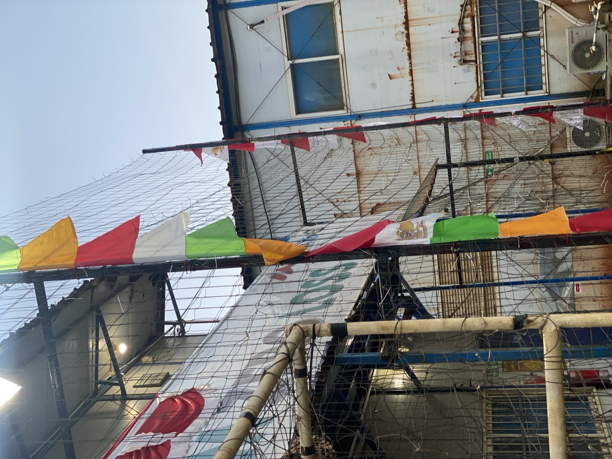 An outdoor, eye-level, medium shot captures a chain-link fence adorned with colorful triangular flags. The flags are arranged vertically, alternating between red, white, green, and yellow. Behind the fence, a dilapidated building with a corrugated metal roof and rusty walls is visible. The building has several windows with blue panes. The scene appears to be an industrial or commercial area, possibly a sports complex or a factory, with an air conditioning unit mounted on the exterior wall of the building. The sky is clear and bright, indicating daytime.