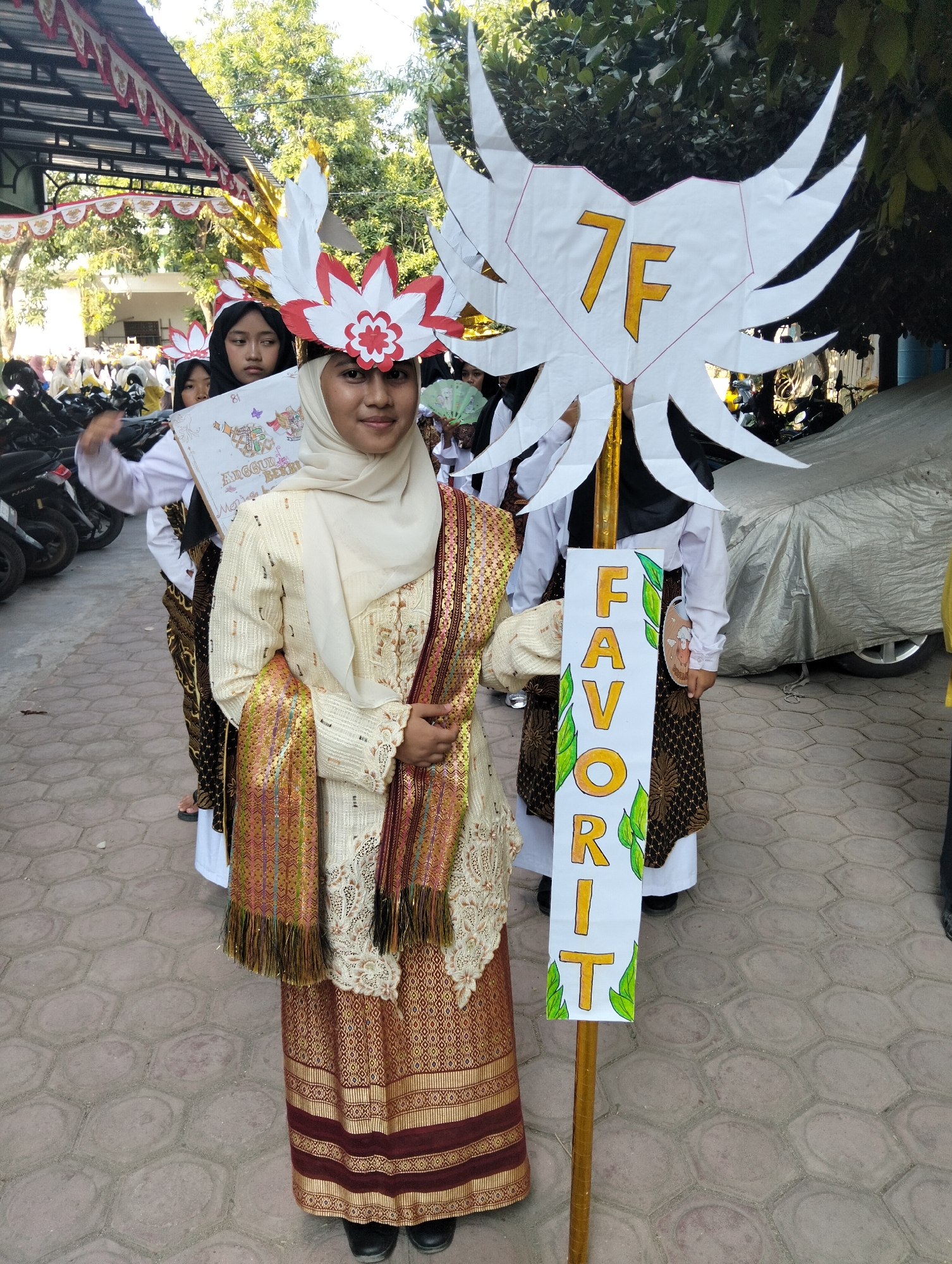 In Kranji, Indonesia, a young Indonesian girl, wearing a traditional cream-colored hijab, a decorative white and red headpiece resembling a flower, and a gold and maroon patterned traditional dress, poses for a photograph. She is holding a sign in her left hand that says "ANGGUL BERRI". In her right hand, she holds a wooden pole with a vertical sign that spells out "FAVORIT" in large yellow letters with green leaf embellishments. Behind her, other girls in similar traditional attire and headpieces are visible, some holding signs and fans. The scene appears to be outdoors, possibly during a parade or cultural event, with motorcycles and a covered car in the background, and trees overhead.