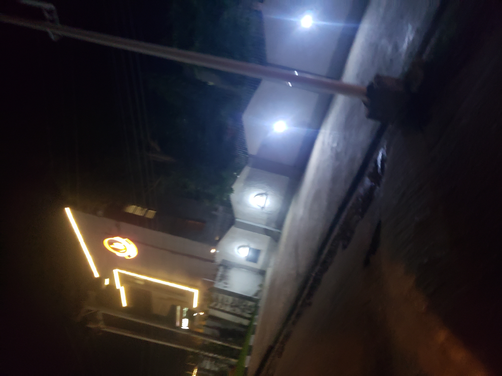 A vertical, night-time photograph captures a building with illuminated signage in Narayi, Nigeria. The building features a prominent neon sign composed of bright yellow lights forming the number "7" and a circular emblem. To the left of the building, a darker structure is visible, also with some lighting. In the foreground, a paved area, likely a street or walkway, is wet and reflects the surrounding lights. A concrete barrier runs along the side of the paved area, and beyond that, a low wall with arched windows is illuminated by bright lights, suggesting interior lighting. A utility pole stands to the right of the frame. The overall impression is a well-lit commercial or residential area at night.