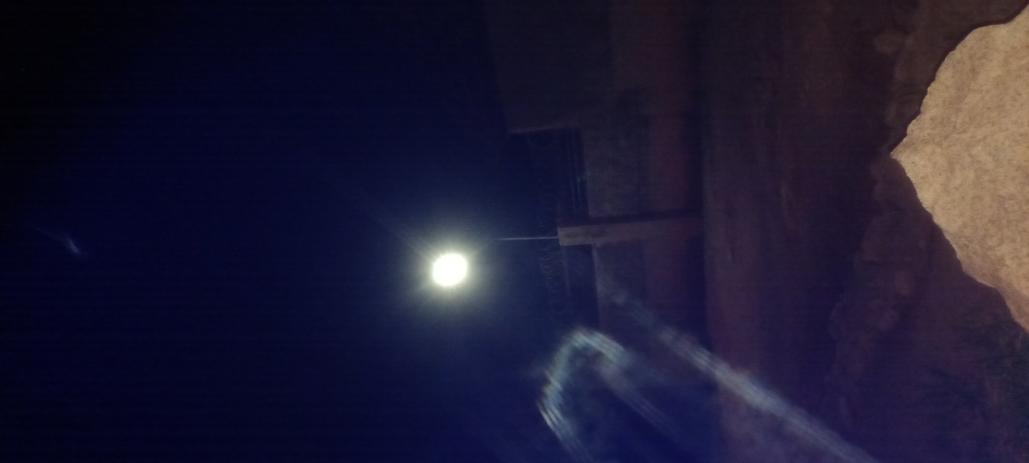 The image depicts a nighttime scene on a street in Kwang, Nigeria. A bright street lamp illuminates the immediate surroundings, casting a strong light upwards and outwards. In the background, a wall topped with barbed wire suggests a perimeter or boundary. The ground appears to be uneven, with some rocky or sandy elements visible in the foreground. There are no people or animals clearly visible in the frame, and the overall atmosphere is one of quiet or deserted urban night. The presence of the street lamp indicates a public or semi-public area within the city.