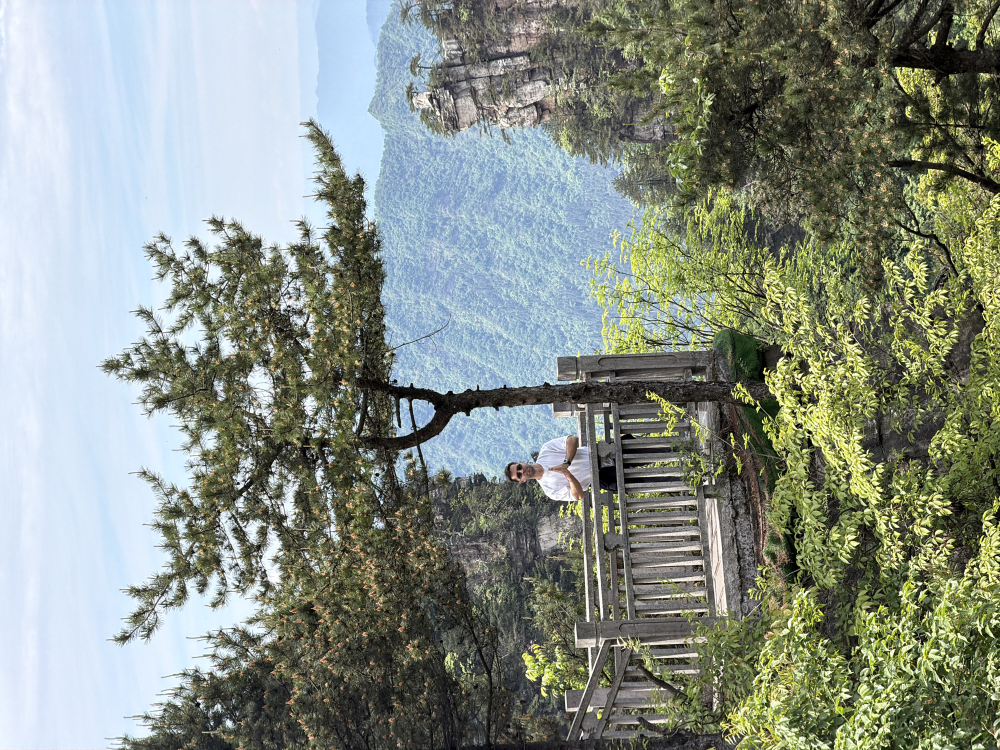 A medium shot of a man leaning on a wooden railing against a backdrop of forested mountains, taken at the Jundiping location in China. The man, wearing a white short-sleeve shirt and sunglasses, has his arms crossed and is looking at the camera. The railing is part of a viewing platform, with the mountains and trees visible behind it. Trees with dense foliage frame the image on both sides. The sky is light blue with a few wisps of clouds. The mountains in the distance are covered in green vegetation, and rocky peaks are visible.