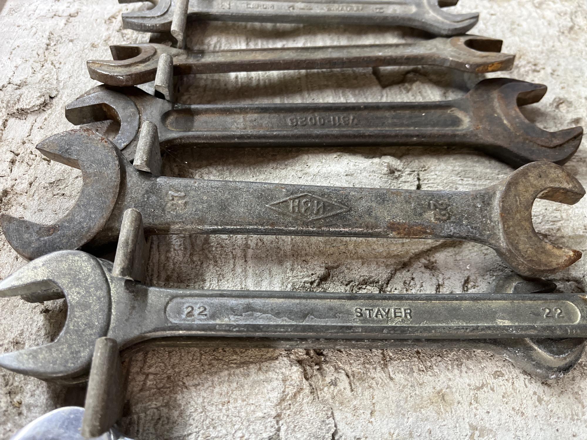 This image displays a collection of vintage, well-used wrenches arranged in a row on a textured, light-colored surface, possibly concrete or stone. The wrenches vary in size and appear to be made of metal, showing signs of rust and wear, indicating their age and extensive use. They are laid out horizontally, with the open ends facing upwards and to the left.
The focus is on the tools themselves, highlighting their industrial nature and history. Some wrenches have markings, with one clearly visible "22" next to the word "STAYER," and another with a diamond-shaped logo containing the letters "HBH." Another wrench shows the numbers "7311-0026." The lighting suggests an indoor or overcast outdoor setting, with soft shadows that don't indicate a specific time of day. The overall impression is one of an organized, yet rustic, display of tools that have seen significant service. No people are visible, and the context of Krasnyye Barrikady, Russia, provides a geographical location without directly influencing the visual content.