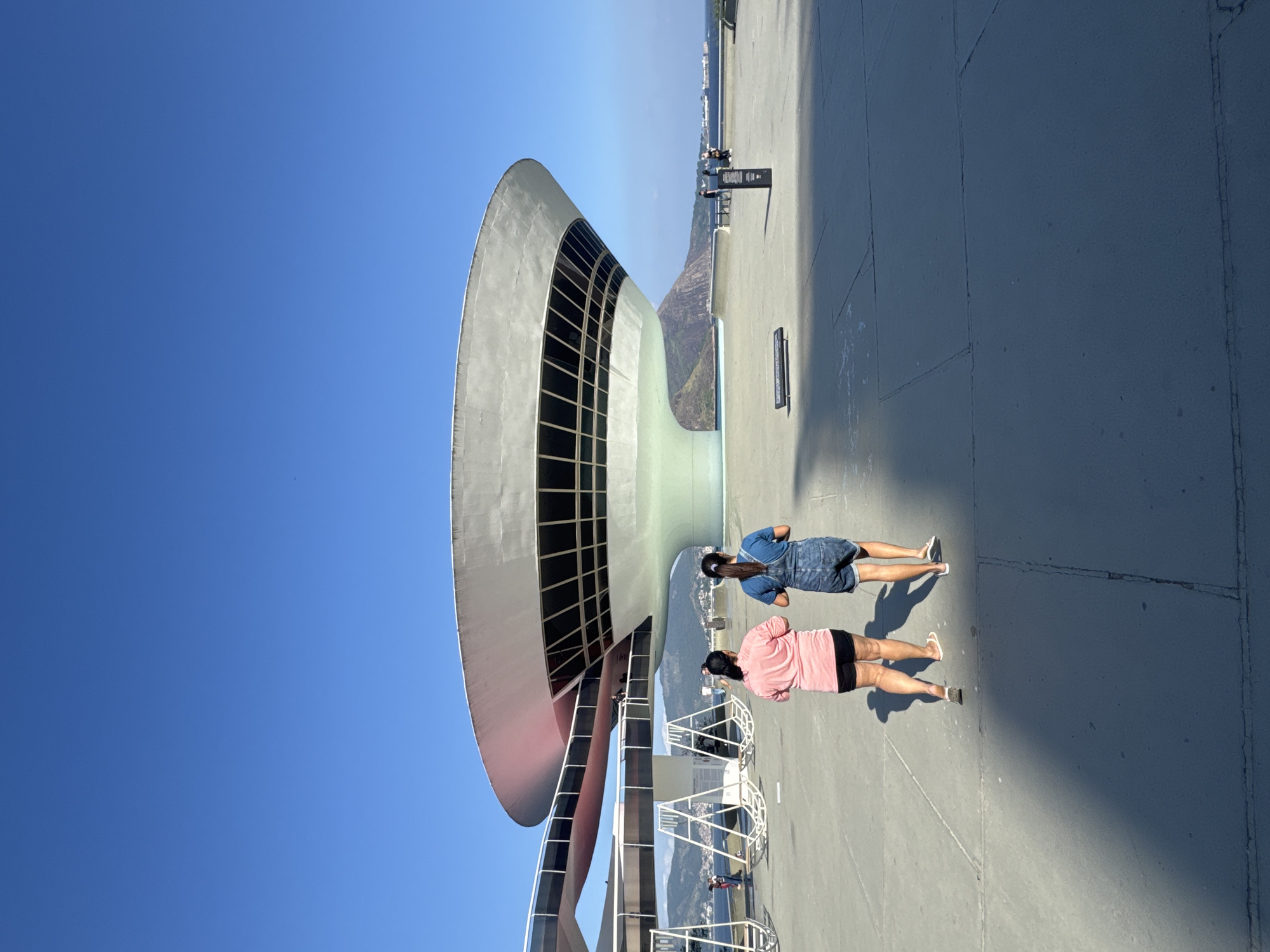 Two young women walk away from the camera on a paved plaza in front of the Niterói Contemporary Art Museum (MAC Niterói) in Niterói, Brazil. The woman on the left wears a pink t-shirt and black shorts. The woman on the right wears a blue t-shirt and denim shorts. The museum, designed by Oscar Niemeyer, is a distinctive saucer-shaped building situated on a peninsula overlooking Guanabara Bay. The sky is clear and blue, and the sun casts long shadows across the plaza. In the background, hills and the bay are visible. Several other people are present on the plaza and near the museum entrance.