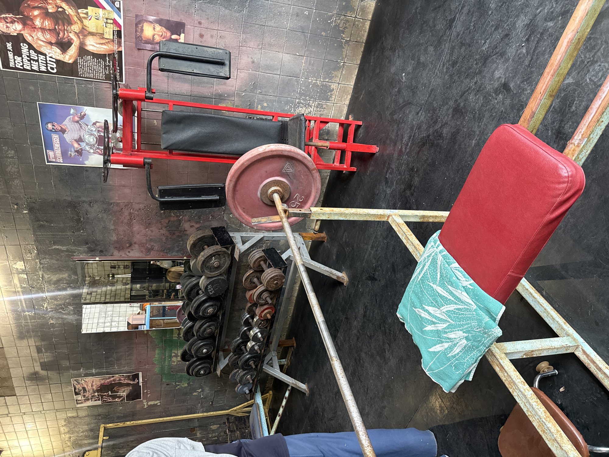 The image depicts the interior of a rustic gym. In the foreground, a red weight bench with a teal towel is visible. To the right, a red weightlifting machine with a black seat is present. A barbell with red weight plates is positioned in front of the machine. The background features shelves stocked with dumbbells of various sizes and weights. Two posters are displayed on the wall: one of Arnold Schwarzenegger on a motorcycle, and another of a muscular man. The gym walls are tiled and appear worn, with some areas showing damage. The floor is black and appears to be made of rubber or a similar material. A person in blue pants is partially visible on the left side of the image, suggesting recent activity. The scene is consistent with a weightlifting facility. This gym is located in Vnukovo, Russia.