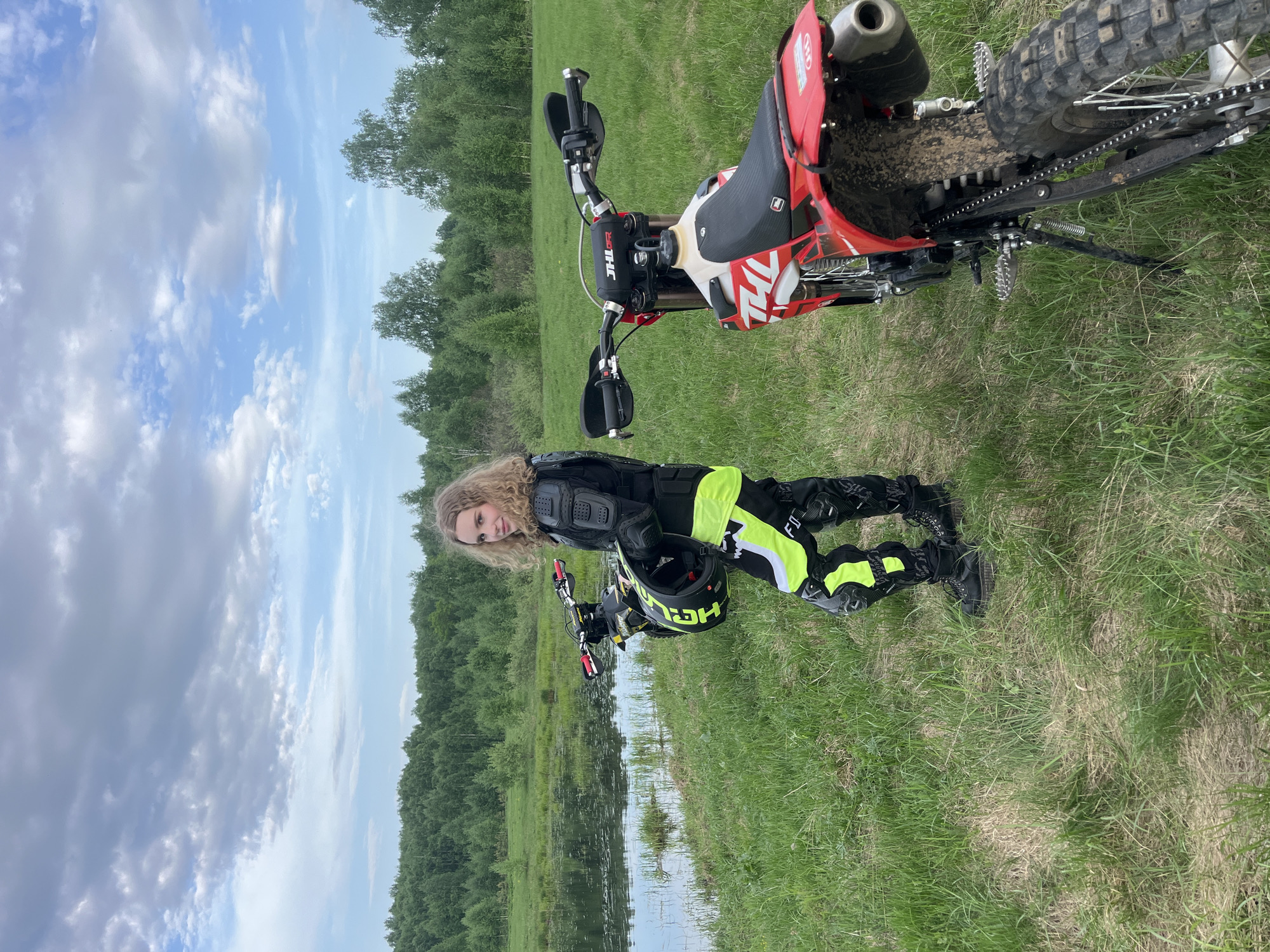 A person with long blond hair is standing outdoors in a grassy field near a red and white dirt bike and a body of water. The person is wearing black and neon yellow protective riding gear, including a chest protector and knee pads. They are holding a black helmet with neon yellow accents. A second dirt bike is partially visible in the background. The location appears to be near a wooded area and a body of water reflecting the trees and cloudy sky. This is taking place in Новочувашский, Russia.