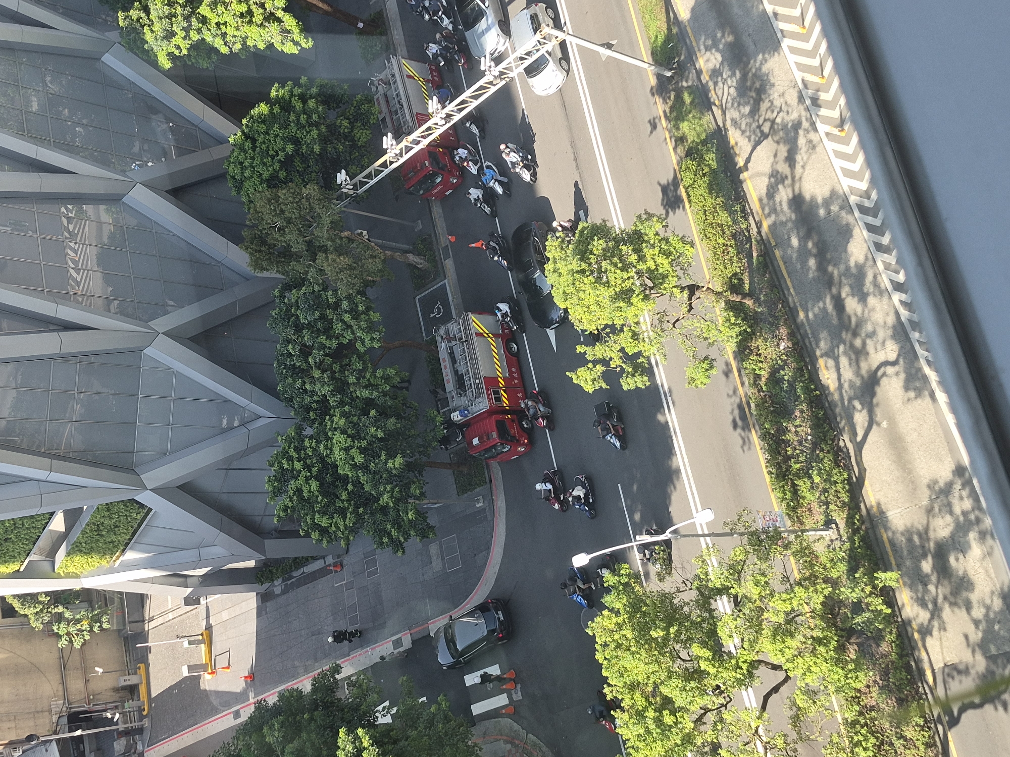 This aerial image captures a street scene in Taipei, Taiwan, characterized by a modern, angular building and a busy road with several vehicles and pedestrians. The building, with its distinctive grey, faceted facade and green vertical accents, dominates the upper portion of the frame. Below, a multi-lane road runs horizontally.
On the road, a red fire truck is parked, its emergency lights appearing to be off. Several motorcycles are in motion and parked along the road. One black sedan is stopped at a pedestrian crossing, where two figures in white are visible. Another silver sedan is also present on the road.
The environment includes mature green trees lining the road and a sidewalk area. The sun casts shadows on the road and surrounding pavement, indicating it is daytime. Traffic cones are also visible, suggesting potential traffic control or construction. The overall impression is of an active urban environment with emergency services present.