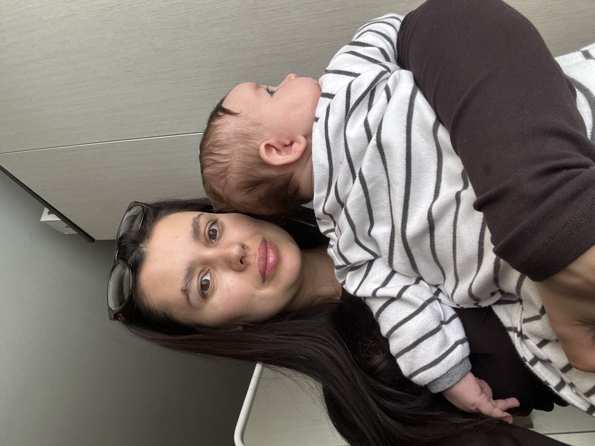 This indoor portrait, taken in Chaikovsky, Russia, features a woman holding a baby. The image is rotated 90 degrees counter-clockwise.
The woman has long, dark brown hair and dark eyes, looking directly at the camera with a neutral to slightly soft expression. She has eyeglasses pushed up onto her head. The baby, with light brown hair, is dressed in a white long-sleeved top with dark stripes and dark brown leggings. The baby is nestled against the woman's shoulder, looking upwards and slightly to the right, away from the camera, with a curious expression visible in profile. The baby's small hand is visible near the woman's hair.
The background consists of plain, light-colored wall or ceiling paneling, featuring a horizontal seam across the upper portion of the frame. The lighting is bright and even, suggesting daytime. No visible text is present in the image.