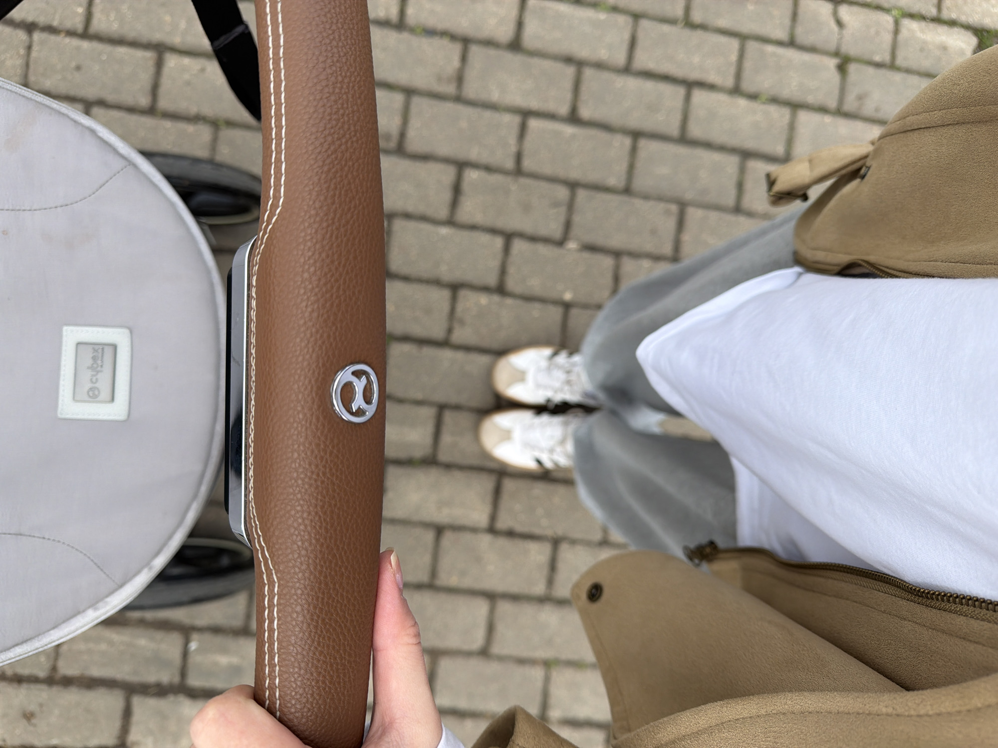 The image presents an overhead, first-person perspective of a person pushing a stroller outdoors. The scene focuses on the stroller's handle, the person's attire, and the paved ground.
In the foreground, a hand, possibly female, grips a brown, textured stroller handle. The handle is accented with white stitching along its length and features a metallic, circular emblem displaying a stylized "C" or similar logo, identifiable as the Cybex brand symbol. To the left, a portion of a light grey fabric stroller or baby carriage is visible, sporting a white tag that clearly displays the text "Cybex" and smaller, less distinct text, possibly "Platinum," below it.
The person is casually dressed in a white t-shirt and grey pants, likely sweatpants. Their feet are clad in light-colored sneakers, predominantly white or beige, with darker accents. On the right side, a beige or tan jacket, appearing to have a suede-like texture, is visible. A zipper and a snap button are notable details on this outerwear. Additionally, a small, matching beige bag or pouch, possibly a fanny pack or an integrated part of the jacket, is partially seen.
The activity taking place is a walk or an outing, as the person is actively holding the stroller handle, suggesting they are pushing it. The setting is an outdoor paved area, likely a sidewalk or pedestrian path, constructed from rectangular grey pavers or bricks. The lighting indicates it is daytime, with soft, diffused light, implying either an overcast day or an evenly lit outdoor environment. The overall impression is a routine, casual moment in Khimki, Russia. No specific emotions are discernible from this vantage point.