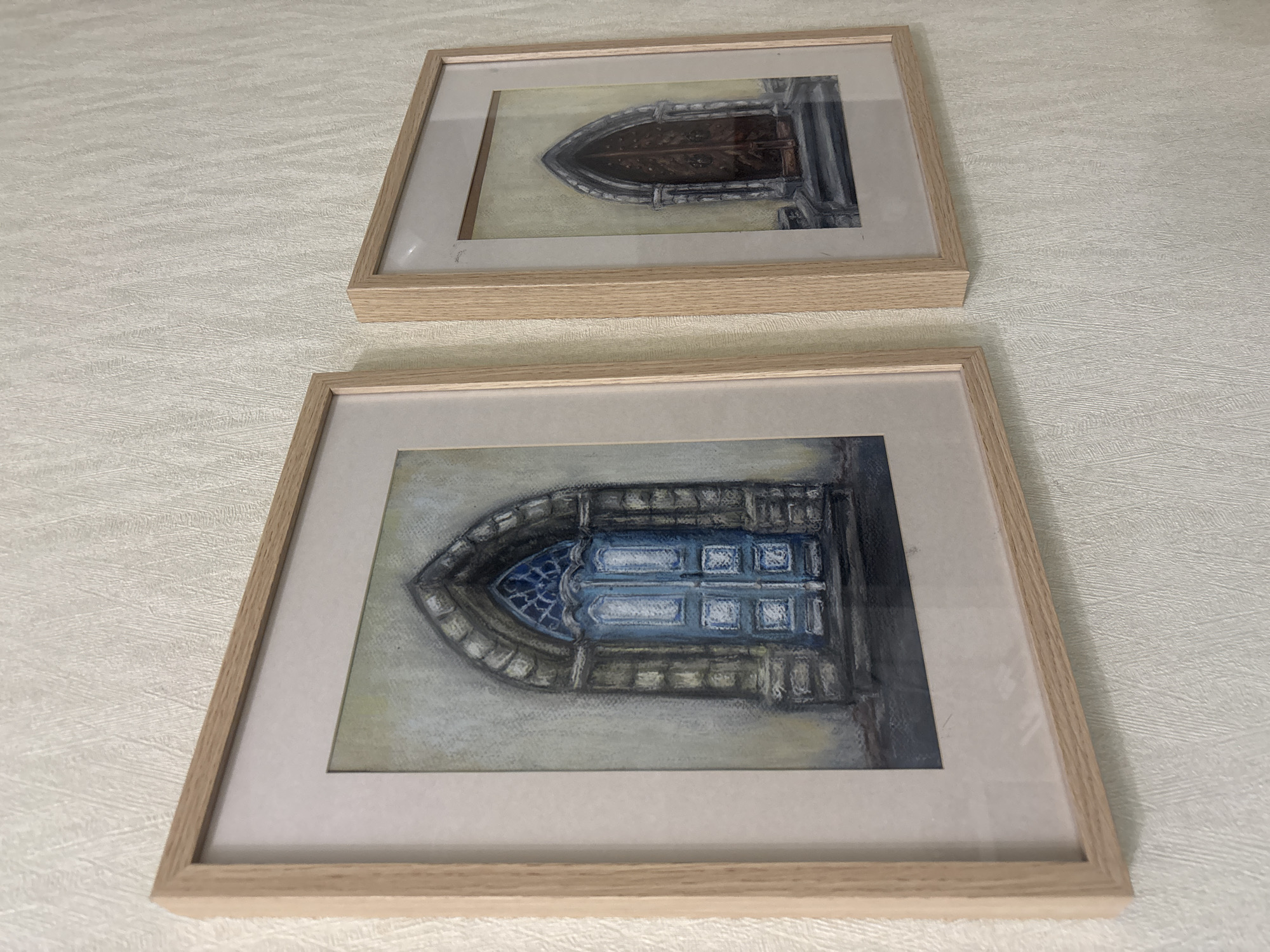 The media file contains two framed pastel drawings of arched doorways. The drawings are displayed on a textured, light-colored surface, possibly a wall or table. Both doorways are depicted with architectural details such as stone surrounds and carved elements.
The top drawing features a dark, ornate wooden door within a gothic-style archway. The lighting suggests an interior view, with shadows cast on the right side of the doorway. The bottom drawing shows a blue door with a smaller, stained-glass window in a similar gothic archway. This drawing also exhibits strong lighting and shadow contrasts, hinting at an exterior or well-lit interior space. The framing for both pieces is simple, with light-colored wooden frames and white matting. The overall impression is one of artistic representation of architectural features, possibly historical or religious buildings, given the style of the doorways. No people, text, specific time of day, weather, or emotional cues are discernible from the drawings or their presentation. The location context provided is Yanino-1, Russia, but there are no visible cues within the image to confirm this.