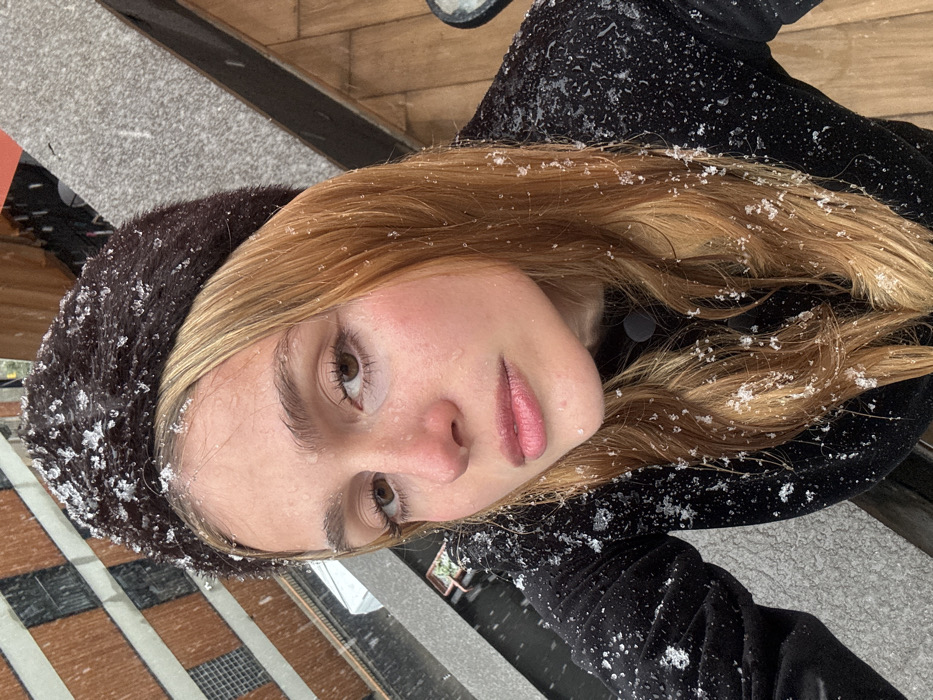 A female subject is depicted in a close-up, high-angle outdoor portrait during snowfall in Unknown, Unknown. She has light brown hair and fair skin, with visible water droplets and melted ice crystals on her face, hair, and dark-colored, long-sleeved garment. A dark brown furry beret is worn on her head, also showing snow accumulation. Her eyes are directed towards the camera. The background reveals urban architectural elements including a building with reddish-brown rectangular panels, a grey textured surface, and a brown wooden surface. A dark railing and a planter containing green foliage are also visible, consistent with an elevated outdoor area such as a balcony or terrace.