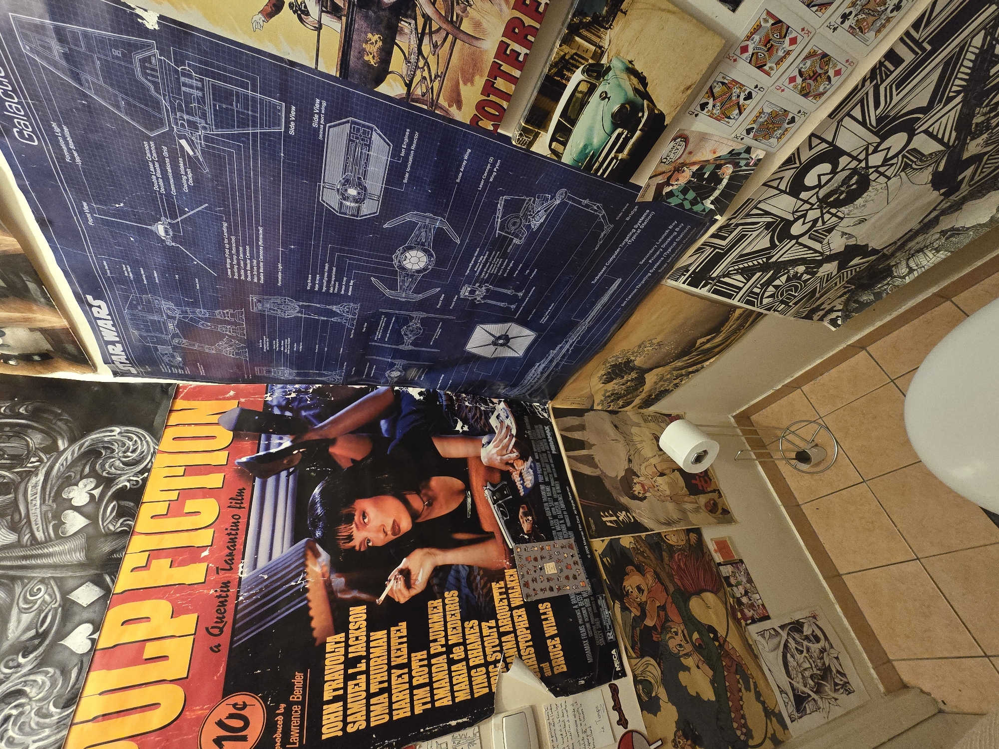 The image shows a collage of posters and other ephemera covering a wall in a room, likely a bathroom, given the presence of a toilet paper holder and toilet in the bottom right corner. The posters include a "Pulp Fiction" movie poster, "Star Wars" blueprints featuring the Millenium Falcon and TIE fighter, and various other images including illustrations, playing cards and comic book art. The posters are taped haphazardly to the wall, creating a dense, layered composition. The environment appears to be a small, cramped space, possibly a bathroom based on the presence of the toilet roll holder and toilet seat. The tiled floor and the perspective of the shot suggest it is a low-angle view taken from within the space. The location in Aubervilliers, France, is not evident in the image.
