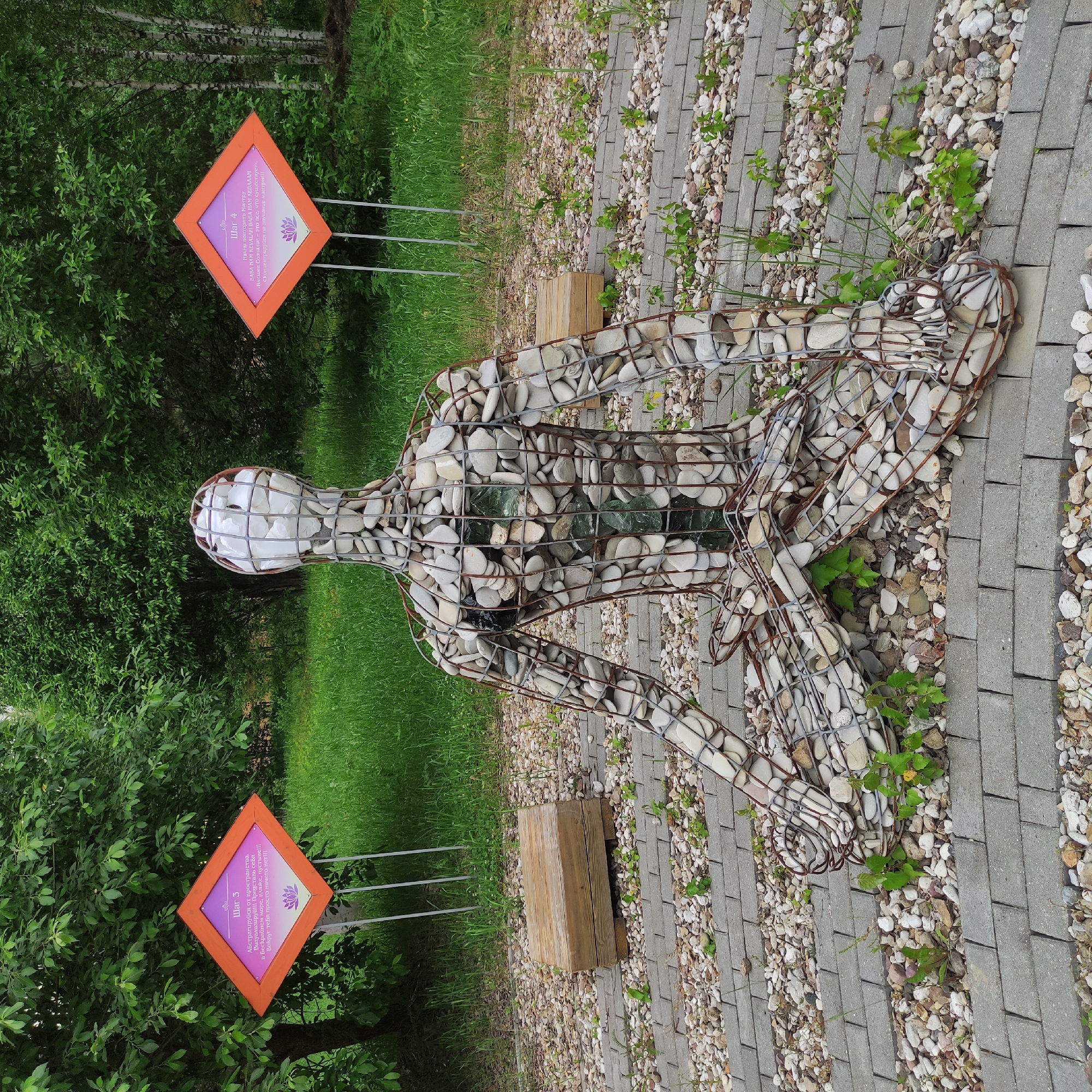 In Zhirnosovo, Russia, a wireframe sculpture of a human figure in a meditative pose is situated outdoors. The sculpture is filled with white and grey river stones, with some green glass elements visible within the chest area. It is placed on a circular path constructed of grey paving stones and smaller river stones. To the left and right of the sculpture are diamond-shaped orange signs with text and a lotus flower image. The left sign, closer to the camera, has Cyrillic text that reads "Шаг 3" (Step 3) and the right sign has "Шаг 4" (Step 4). Behind the sculpture, a lush green forest with trees and tall grass forms the backdrop. In the middle ground, a wooden crate sits on the stone path to the left of the sculpture.