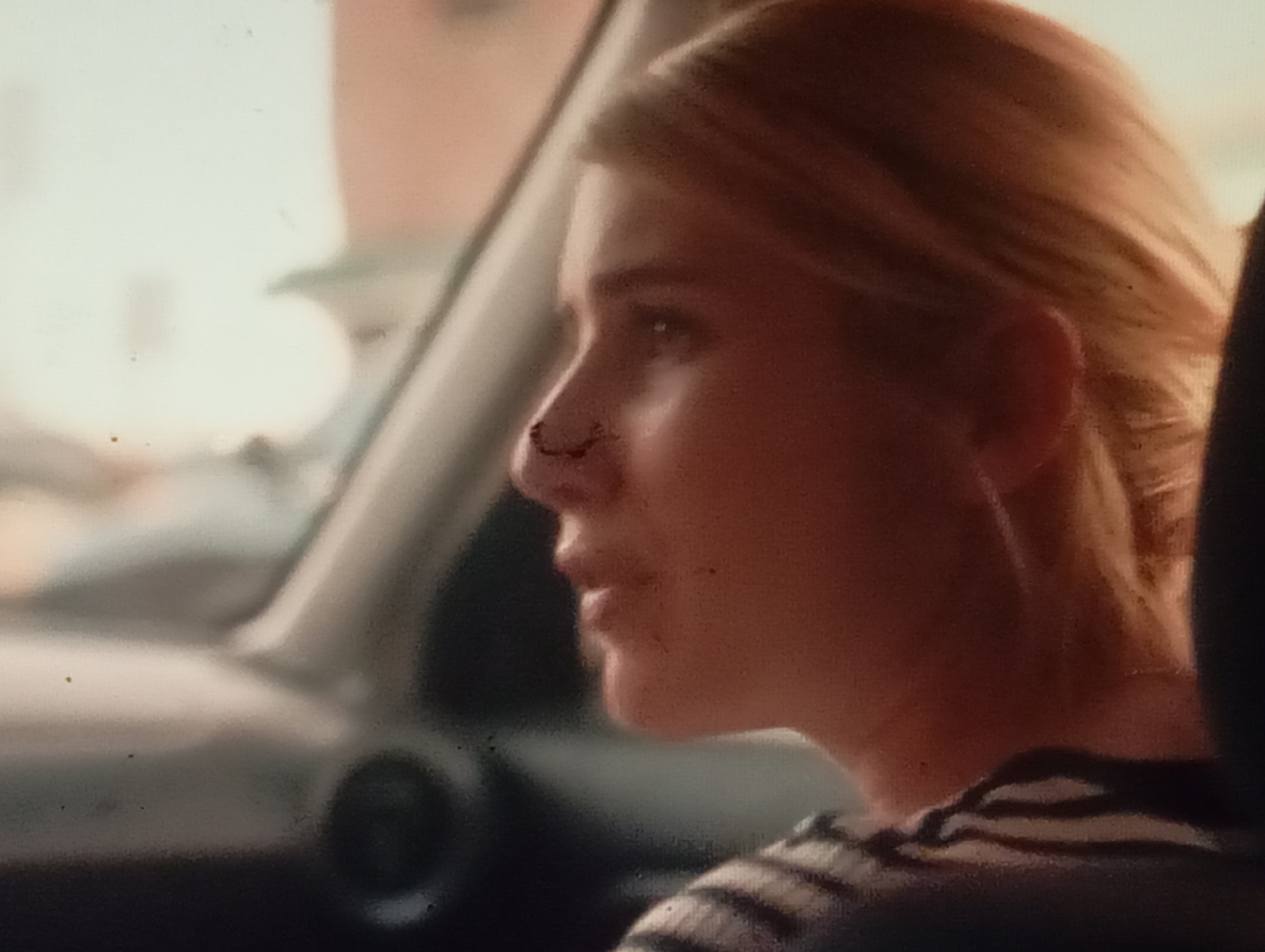 This is a close-up, side-profile shot of a young Caucasian woman with blonde hair, inside a vehicle. The woman is looking out of the vehicle's window. Her expression appears neutral, perhaps a bit weary or thoughtful. There is a small mark or wound on her nose. She is wearing a striped shirt. The setting appears to be a daytime scene, possibly during daylight hours, given the bright, diffused light entering the vehicle. The interior of the car is visible, including part of the dashboard and window frame. The background outside the window is blurred, suggesting motion or a shallow depth of field, but hints at an urban or suburban environment with buildings and possibly some greenery. The image quality is somewhat grainy, which may be due to low light conditions or the camera used. There is no visible text in the image. The location context provided is Sofiivska Borshchahivka, Ukraine, which aligns with a common urban or suburban setting.