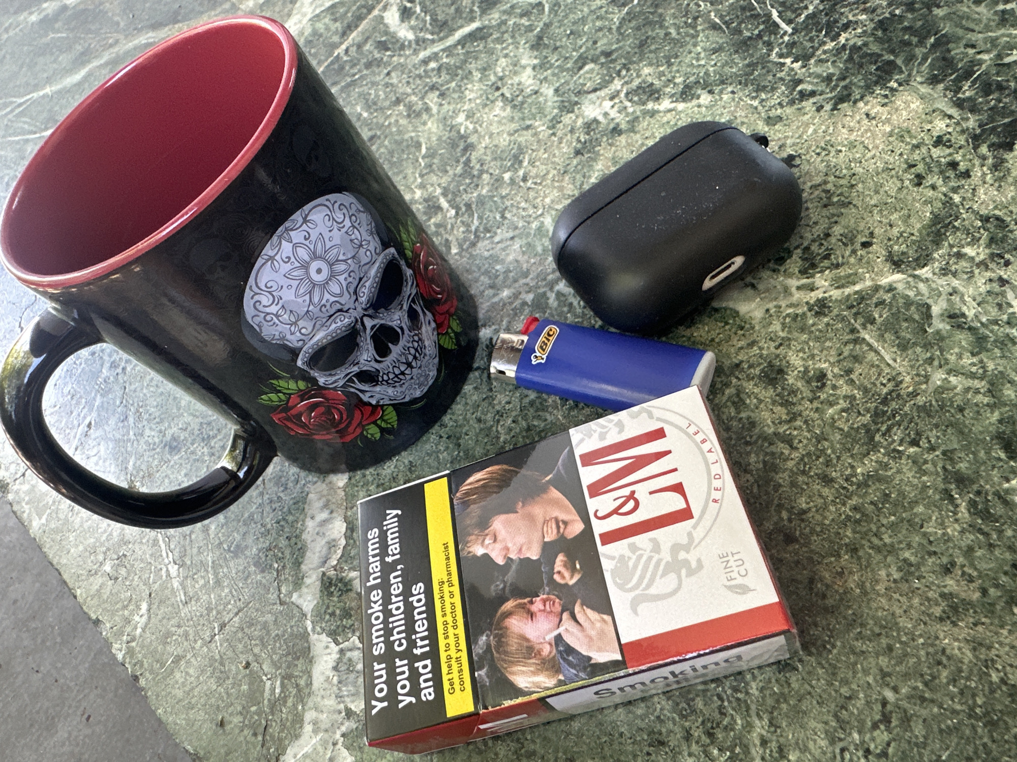 An overhead medium shot shows a collection of items on a marble-patterned surface. In the upper right quadrant, a black mug with a red interior is tilted to the left. The exterior of the mug features a large, detailed sugar skull design with roses. To the left of the mug, a pack of L&M Red Label Fine Cut cigarettes is positioned diagonally. The cigarette pack has a warning label that reads "Your smoke harms your children, family and friends" with a yellow band below that states "Get help to stop smoking: consult your doctor or pharmacist." The front of the pack displays a graphic image of a crying child and an adult holding a cigarette. Next to the cigarette pack, a blue BIC lighter lies horizontally. To the right of the lighter is a black charging case, possibly for earbuds. The marble surface appears to be an outdoor table.