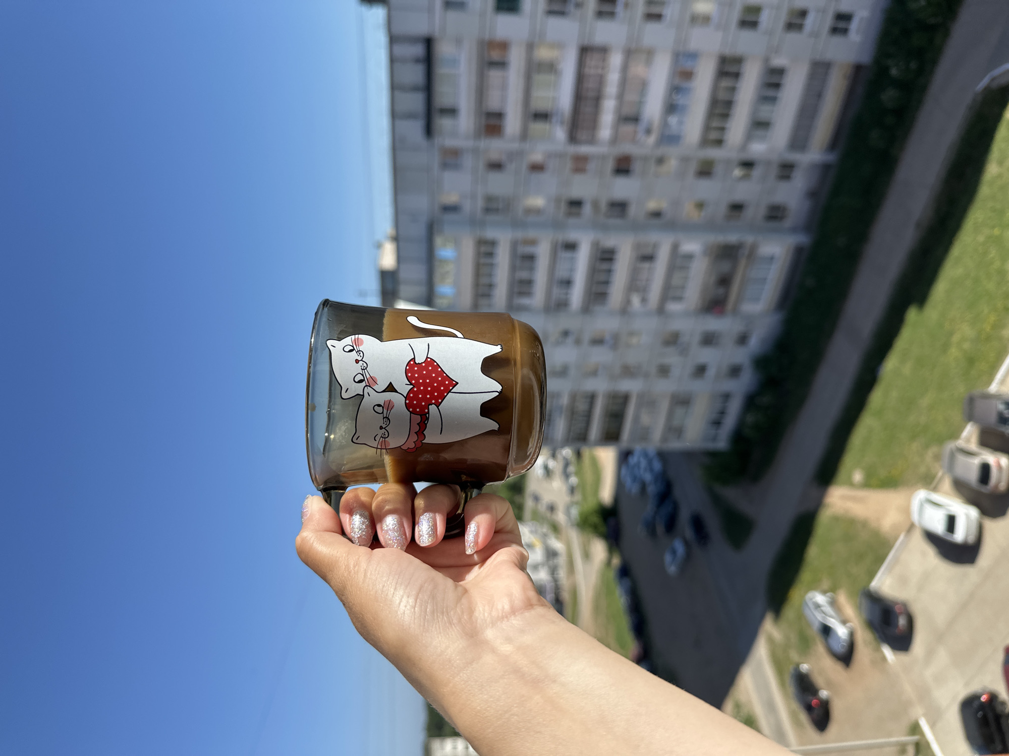 A person's hand holds a glass mug against a backdrop of a bright blue sky and a multistory apartment building, typical of urban Russia. The mug features a cartoon graphic of two cats in love. The building behind the mug has numerous windows and balconies, suggesting a densely populated residential area. Below the building, parked cars are visible, indicating the presence of parking spaces. The manicured nails suggest an effort to present an attractive personal appearance. The specific location is Chaikovsky, Russia.