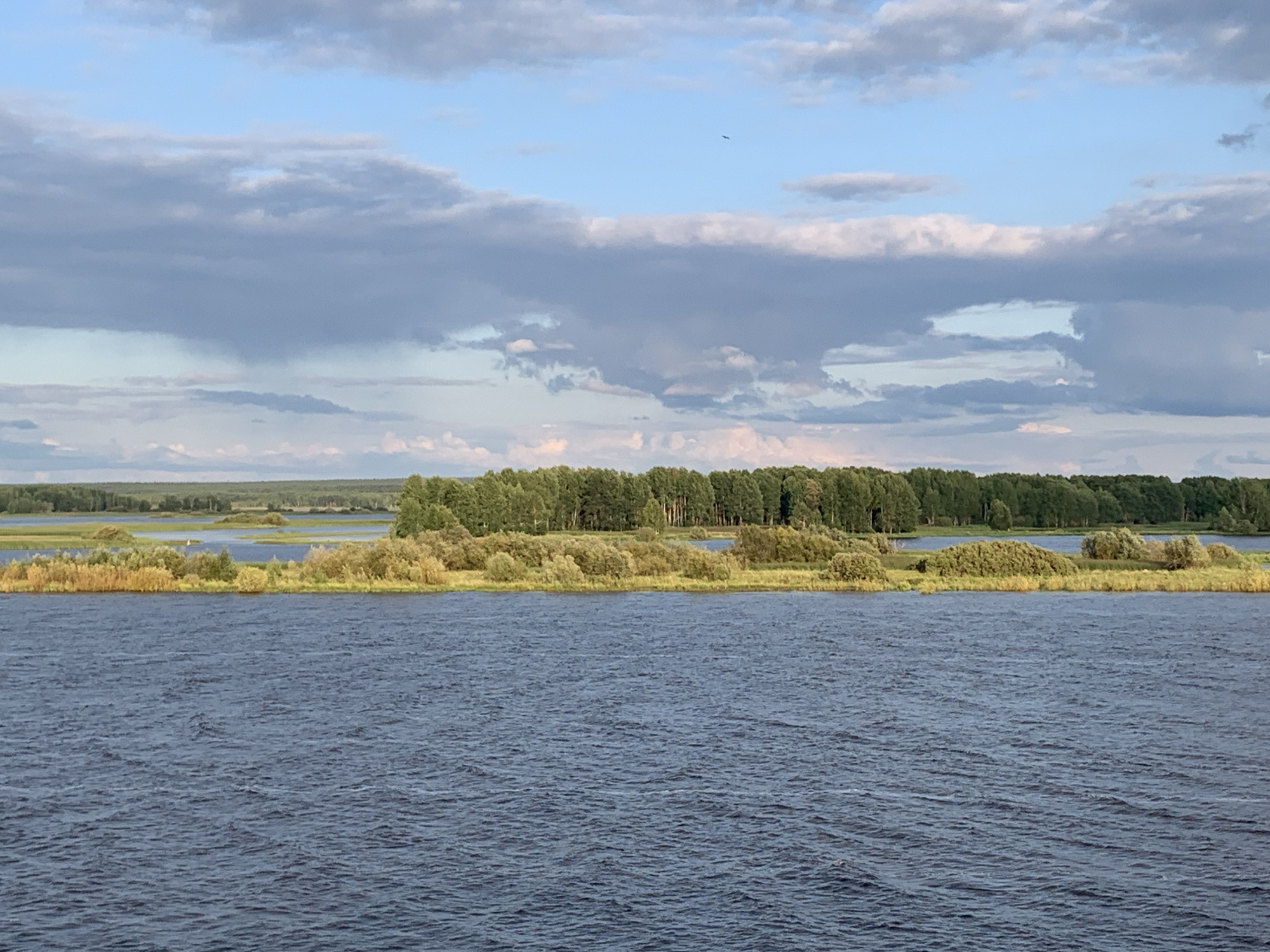 The image depicts a wide, dark blue river in the foreground, with ripples indicating a gentle current. The middle ground features a lush green treeline bordering a body of water dotted with islands covered in vegetation and trees. The far background shows a distant, hazy horizon under a sky filled with dramatic clouds. A solitary bird is visible in the upper right quadrant of the sky. The scene appears to be a natural landscape, possibly a river or lake in a rural or undeveloped area. The location is identified as Berezniki, Russia, suggesting a natural water body within or near the city.