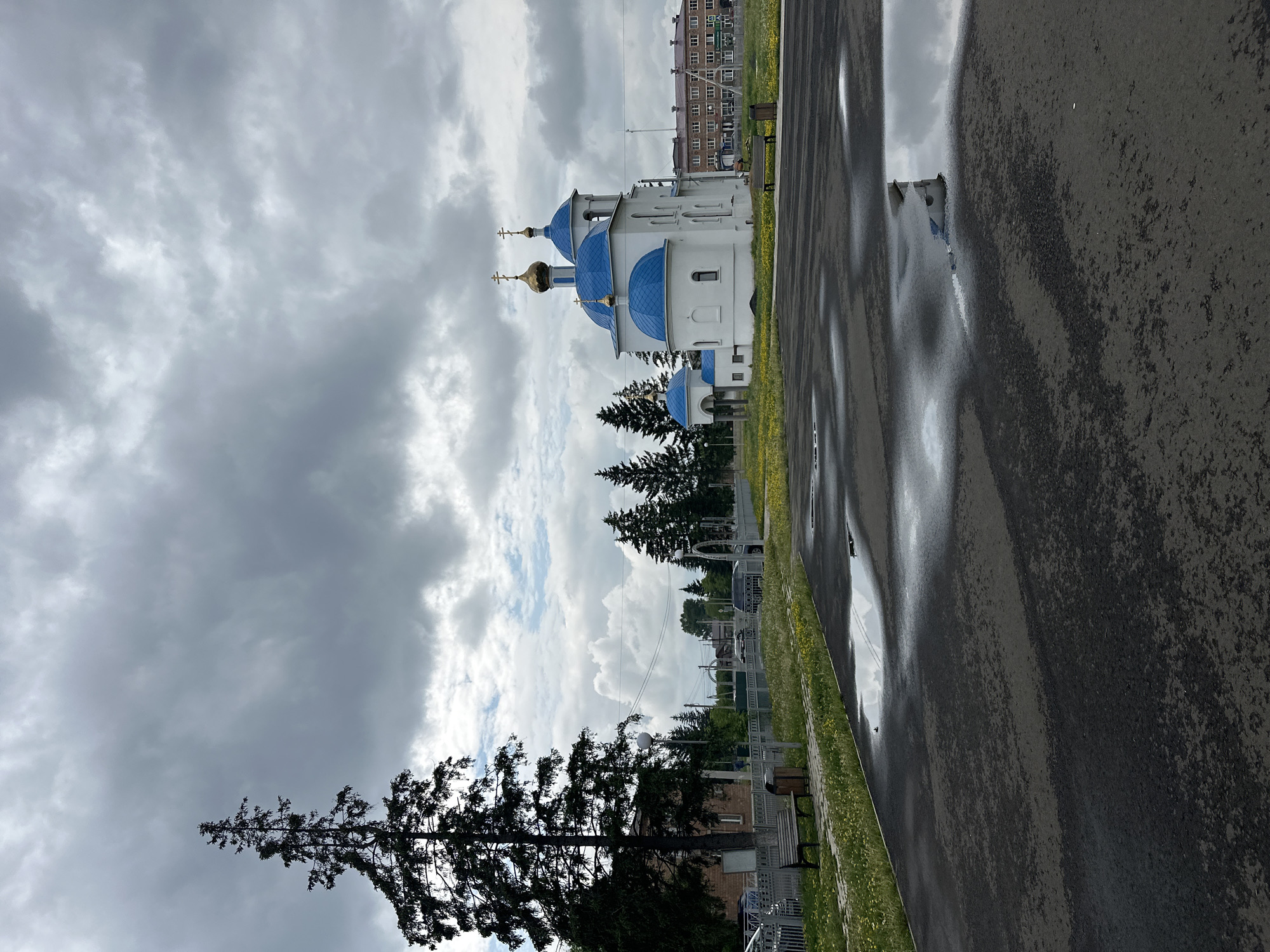 The image depicts an outdoor scene in the city of Dzerzhinskoye, Russia. In the foreground, a wet asphalt surface is visible, with several puddles reflecting the overcast sky and some structures. To the left, a tall, dark evergreen tree dominates the frame. Further back, a white building with bright blue domes and golden crosses, characteristic of an Orthodox church, stands prominently. Adjacent to the church, a long, multi-story brick building can be seen. The environment includes a grassy area with scattered yellow flowers, a white fence, a park bench, and a trash receptacle. Several parked vehicles are also visible near the fence. The sky is heavily clouded with dramatic, gray cloud formations.