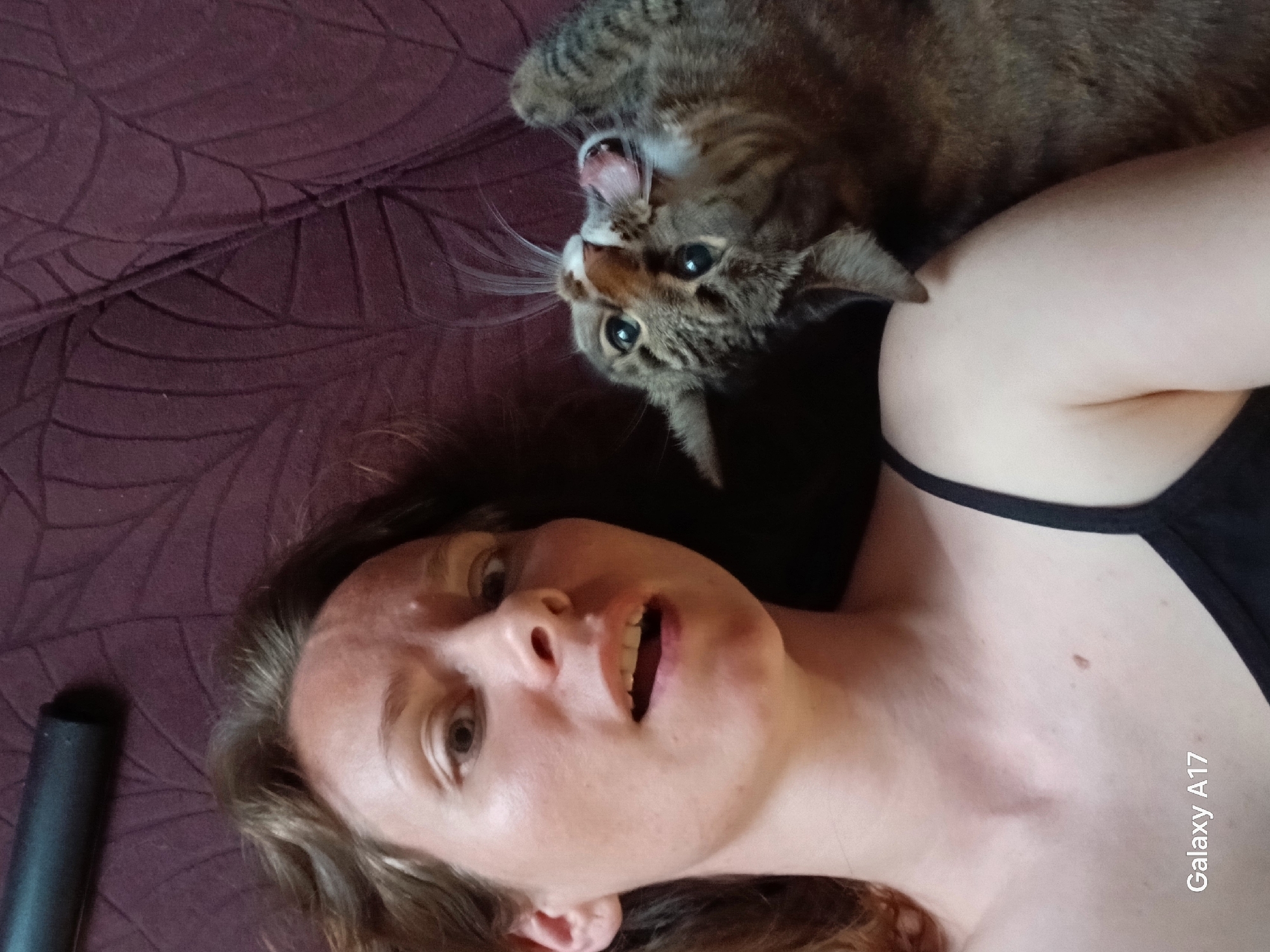 A woman lies on a patterned aubergine-colored surface, possibly a bed or sofa. She is wearing a black tank top. Her head is slightly tilted, and she is looking at the camera with her mouth open. A tabby cat is draped over her shoulder, its mouth open in a possible yawn or hiss. A dark object, possibly a remote, is visible in the lower left corner. The location is unknown, and no features indicate it is Yekaterinburg, Russia. The image also has a watermark stating the picture was taken on a "Galaxy A17".