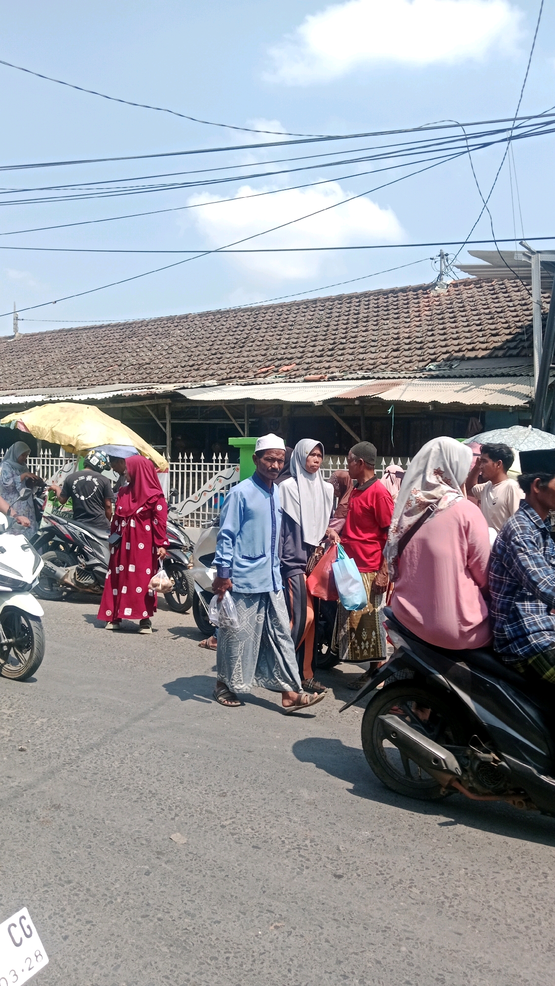 In Waru, Indonesia, a street scene unfolds under a bright, partly cloudy sky. A group of people, predominantly dressed in traditional Islamic attire, are gathered near a row of shops with tiled roofs. The foreground shows a paved road with several motorcycles.
A man wearing a blue long-sleeved shirt and a white skullcap, with a patterned sarong, is in the center, holding a plastic bag. To his left, a woman in a red traditional dress with white polka dots stands near a motorcycle. Another woman in a light grey hijab stands beside the man. To the right, a person wearing a pink hijab is seated on a motorcycle. Further back, other individuals are visible, some holding shopping bags, and one person in a red shirt. A white motorcycle is parked on the left. A yellow umbrella provides shade for part of the area behind the people. The overall atmosphere suggests a bustling market or shopping area.