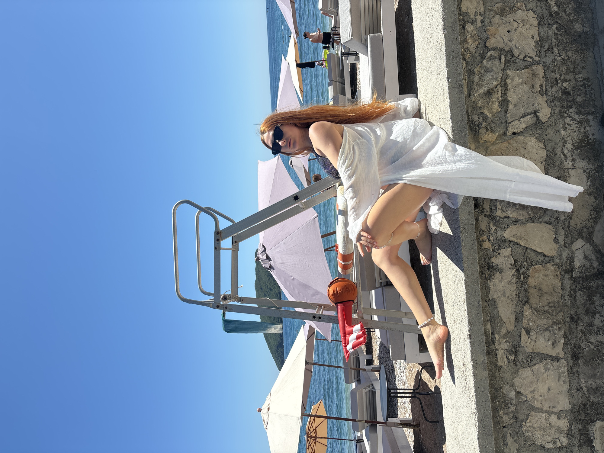 This image depicts a sunny seaside scene in Budva, Montenegro, dominated by a young woman posing on a stone ledge. The woman has long, reddish-brown hair and is wearing dark sunglasses and a flowing white cover-up or dress over a dark swimsuit. She is seated or crouching on a light-colored stone wall that forms a promenade or pier, with her bare feet and a pearl ankle bracelet visible.
Behind and to her left, a metal ladder structure, possibly part of a diving platform or a lifeguard stand, rises from the water's edge. An orange and red-striped lifebuoy is hanging from this structure. The clear blue sea stretches out to the left and into the background, where several light-colored beach umbrellas (some pinkish, some beige) and white sun loungers are arranged along the shoreline. Other people can be seen relaxing on these loungers or by the water, enjoying the bright weather. Distant green hills or mountains are visible across the water, adding to the scenic coastal landscape.
The sky is a brilliant, cloudless blue, indicating clear weather. The strong sunlight casts sharp shadows, suggesting it is midday or early afternoon. The overall atmosphere conveys a sense of relaxation and leisure at a popular beach or resort location. No visible text is present in the image.