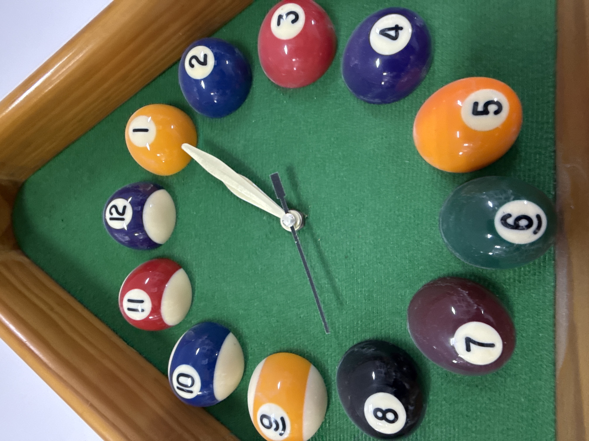 A top-down shot shows a clock made from a miniature pool table with billiard balls marking the hours. The table has a wooden frame and green felt surface. The balls are arranged in a circle, with the number "1" ball at the 1 o'clock position, "2" at 2 o'clock, and so on up to "12". Clock hands protrude from the center. The background around the pool table clock is a plain white surface. There is no indication in the image that it is located in Chaikovsky, Russia.