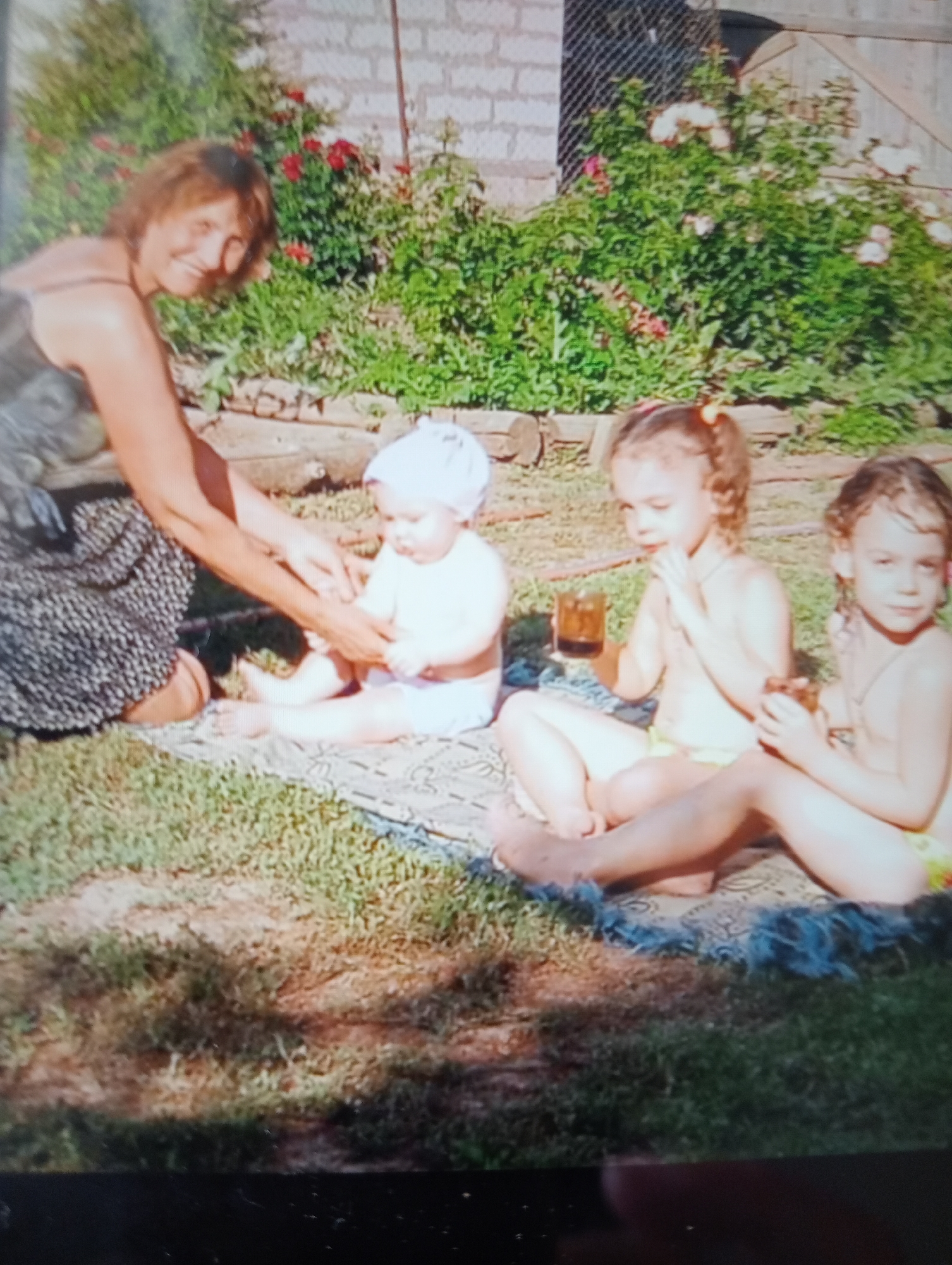 An outdoor, eye-level, medium shot captures a woman and three young children on a patterned blanket in the grass. To the left, a woman with short brown hair smiles as she interacts with a baby in the center. The baby, wearing a light-colored hat and diaper, sits on the blanket, facing away from the viewer. To the right of the baby are two young girls. The girl on the left has curly brown hair and is wearing a yellow swimsuit bottom. She holds a glass in her right hand. Next to her, the girl on the right, also with brown hair, wears a similar yellow swimsuit bottom and holds a glass in her left hand. Both girls are facing the viewer. Behind them, a rose bush with pink and red flowers is visible against a brick wall. The ground is covered with green grass and some dry patches.
