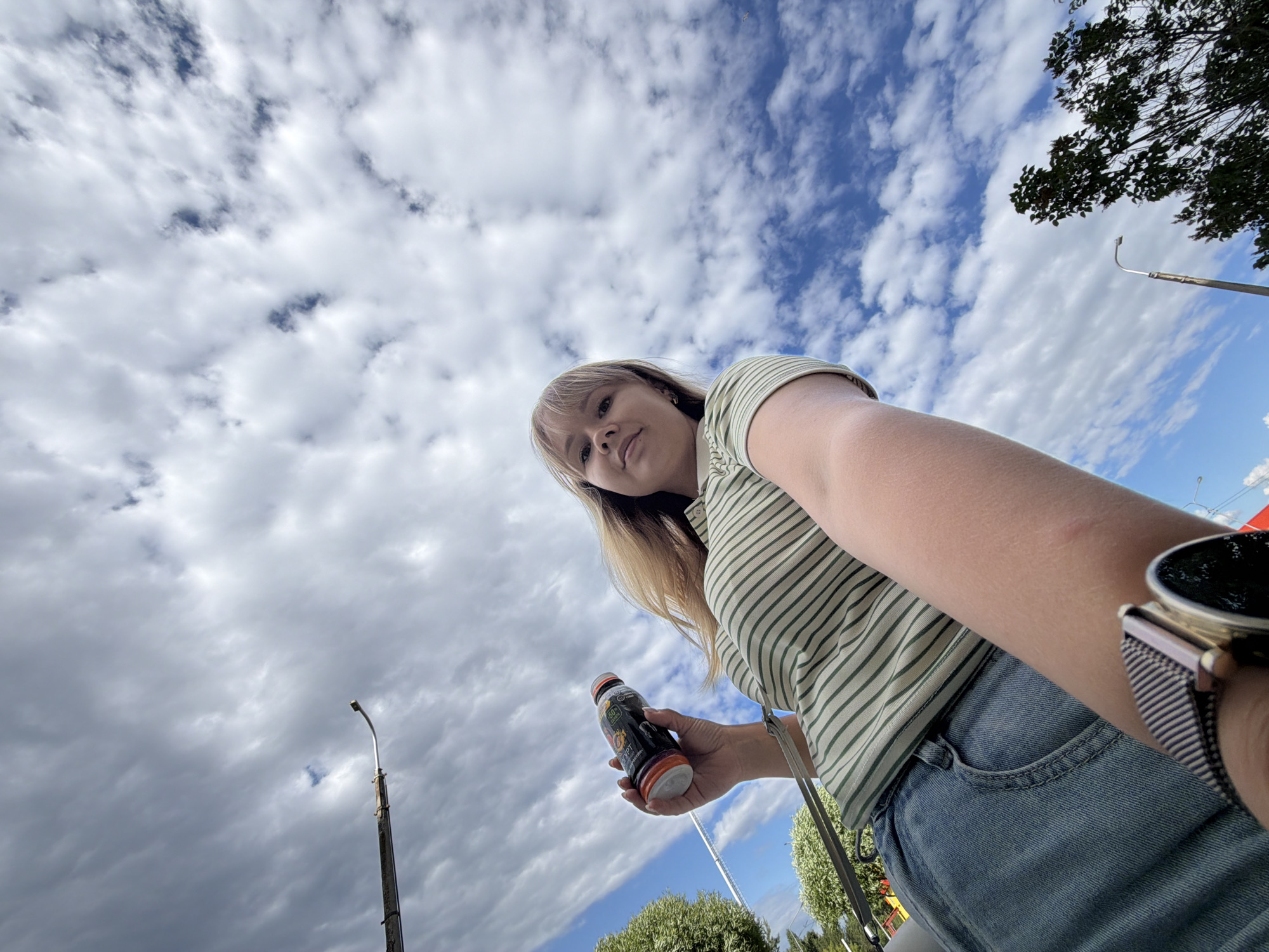 A low-angle, wide shot captures a young woman holding a beverage bottle while standing outdoors, with a cloudy blue sky dominating the background. She is wearing a green and white striped short-sleeved shirt and blue denim jeans. The woman has blonde hair and is looking up towards the camera. Her right arm is extended, holding a dark-colored bottle with an orange cap and label. A metal watch with a striped band is visible on her left wrist. In the background, trees and a lamppost are present. The image appears to have been taken in an urban park or outdoor area. There is no specific information in the image to identify the location as Chaikovsky, Russia.