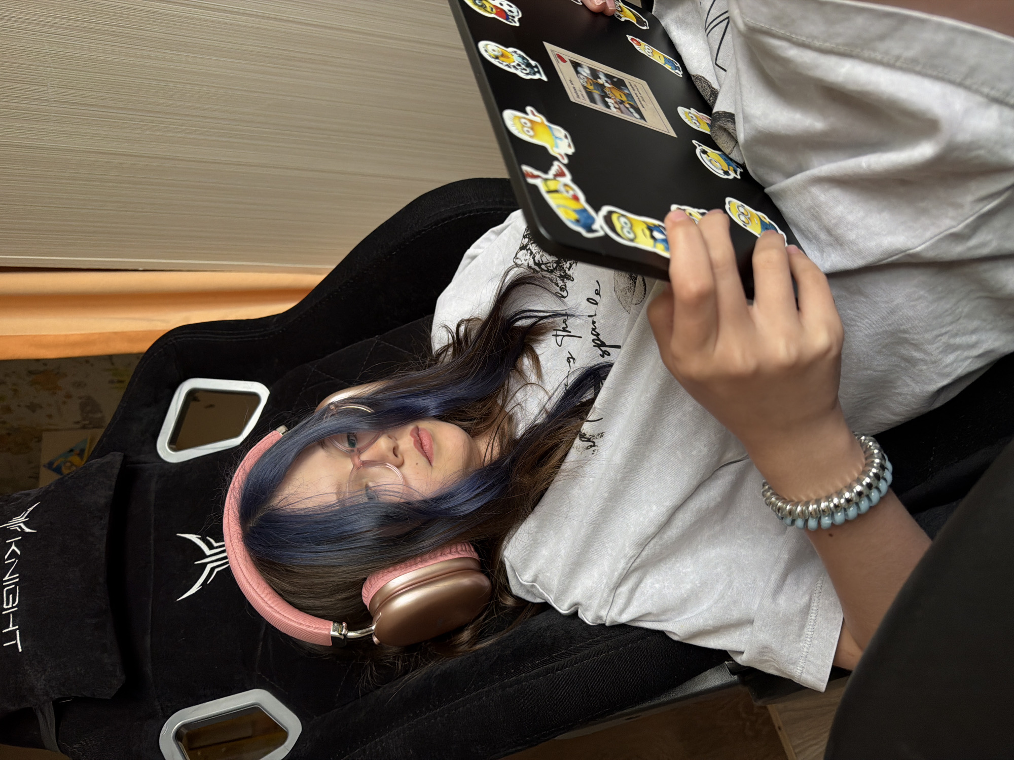 A young person with dark hair featuring blue highlights, wearing round glasses and rose gold over-ear headphones, is depicted lying on their back in a black gaming chair. They are dressed in a light grey t-shirt with partially obscured cursive text and dark pants. A bracelet composed of silver-colored spiral rings and light blue beads adorns their right wrist.
The individual is holding a black laptop or tablet that is extensively decorated with various "Minions" character stickers and a central sticker resembling a playing card. Their hands are positioned on the device, suggesting engagement with its content. The gaming chair is upholstered in black fabric and features the white embroidered word "KNIGHT" along with a stylized logo. In the background, a light-colored, horizontally textured wall and a warm-toned wooden surface are visible, indicating an indoor environment. The person's expression appears neutral and focused on the screen of the device. This scene is situated in Birobidzhan, Russia.