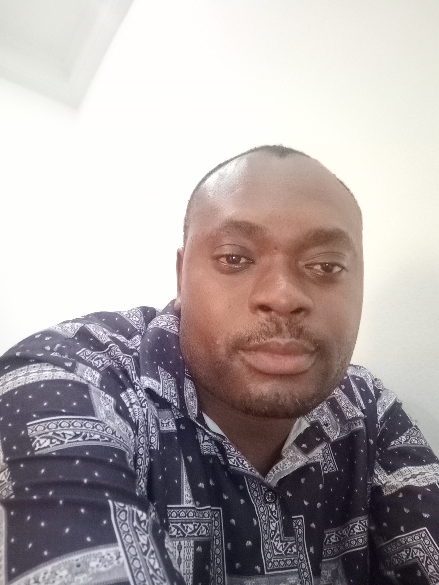 A medium close-up shot features a man with dark skin and short black hair. The man is facing forward and his gaze is directed towards the camera. He is wearing a dark blue button-down shirt patterned with white designs. The background is a plain white wall. There is no information to verify that the photo was taken in Gwarinpa, Nigeria.