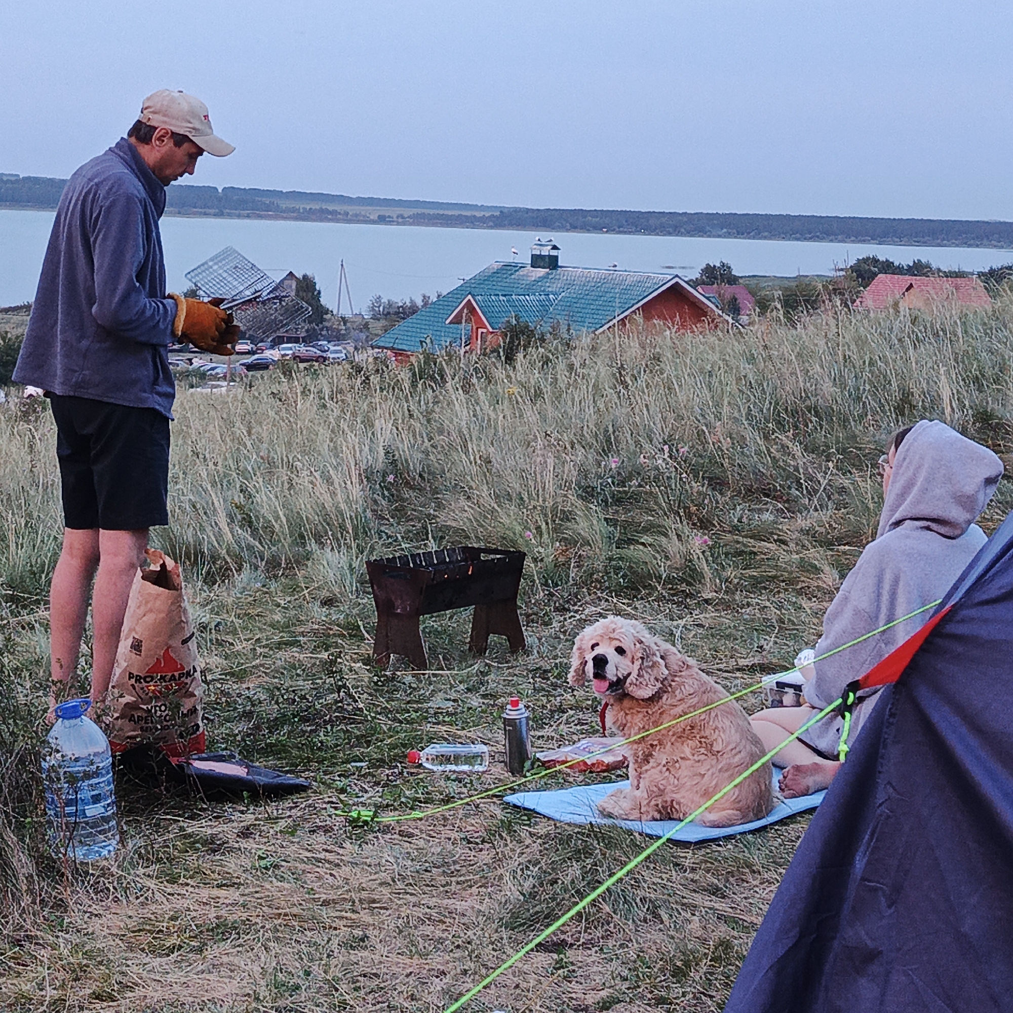 In the city of Yangi-Turmush, Russia, an outdoor camping scene unfolds against a backdrop of a serene body of water and distant trees. The sky is overcast, suggesting a mild, possibly humid, climate. In the foreground, a man wearing a cap and casual clothing is preparing for a barbecue, holding a grilling rack. Beside him stands a large bag of charcoal, and a water bottle is positioned nearby. A metal barbecue grill is set up, with embers glowing within, indicating it is ready for use. A light-colored dog is seated on a blue mat, looking towards the camera with its mouth slightly open. To the right, a person in a hooded sweatshirt sits next to a tent. The ground is covered in dry grass and a few scattered wildflowers. In the middle ground, several houses with red roofs are visible, and a parking lot with several cars suggests proximity to a populated area.
