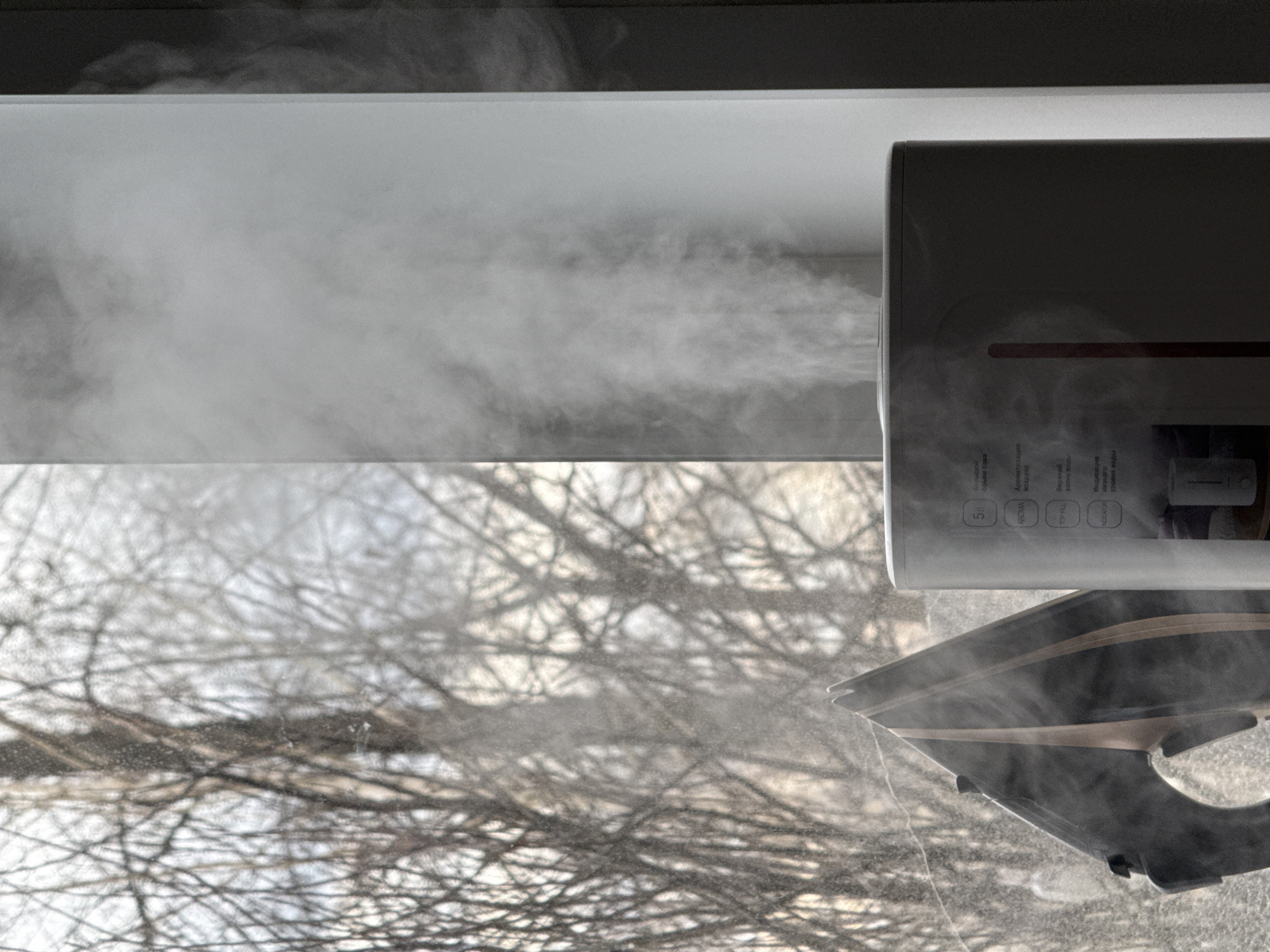 The image depicts a humidifier positioned near a window, with steam visibly emanating from it. Outside the window, bare tree branches are visible. The controls of the humidifier are labeled in Russian, with terms such as "AROMA," "TOP FILL," and "INDICATOR" (rendered in Russian). The text also indicates "Большой объем Сака", which translates to "Large volume Saka." This location could possibly be in or near the city of Chaikovsky, Russia, due to the Russian text on the humidifier.