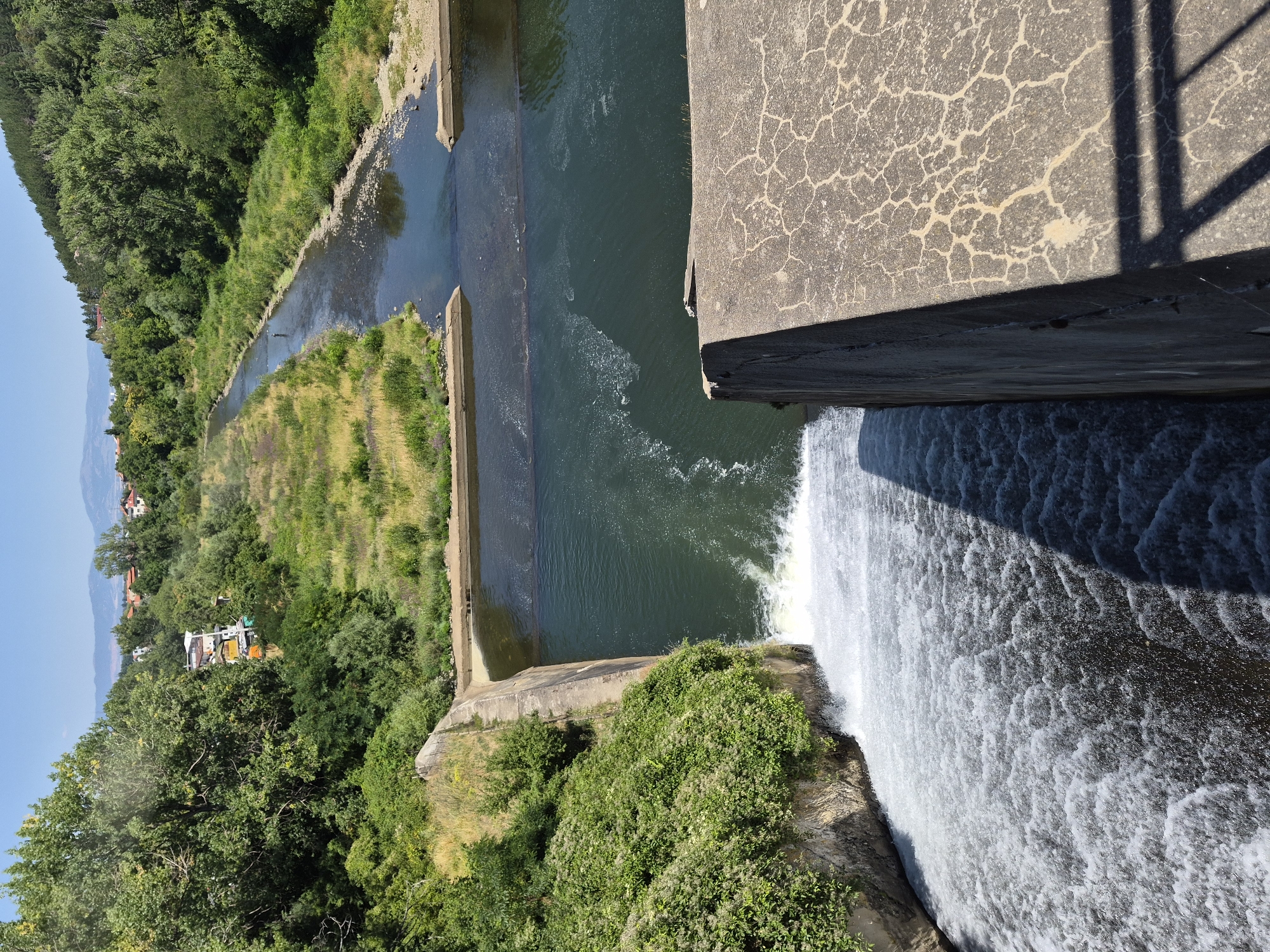 The image shows a section of the Pancharevo Lake dam in Pancharevo, Bulgaria. A dam structure, made of concrete, is visible in the foreground, with water cascading over its edge. The water is turbulent and foamy as it falls. Beyond the dam, a body of water, likely the lake, is visible, with a river flowing into it. The riverbanks are lined with lush green vegetation, including trees and grass. In the distance, hills covered in trees can be seen, and a few buildings are nestled amongst the greenery. The sky is clear and blue, indicating a sunny day.
