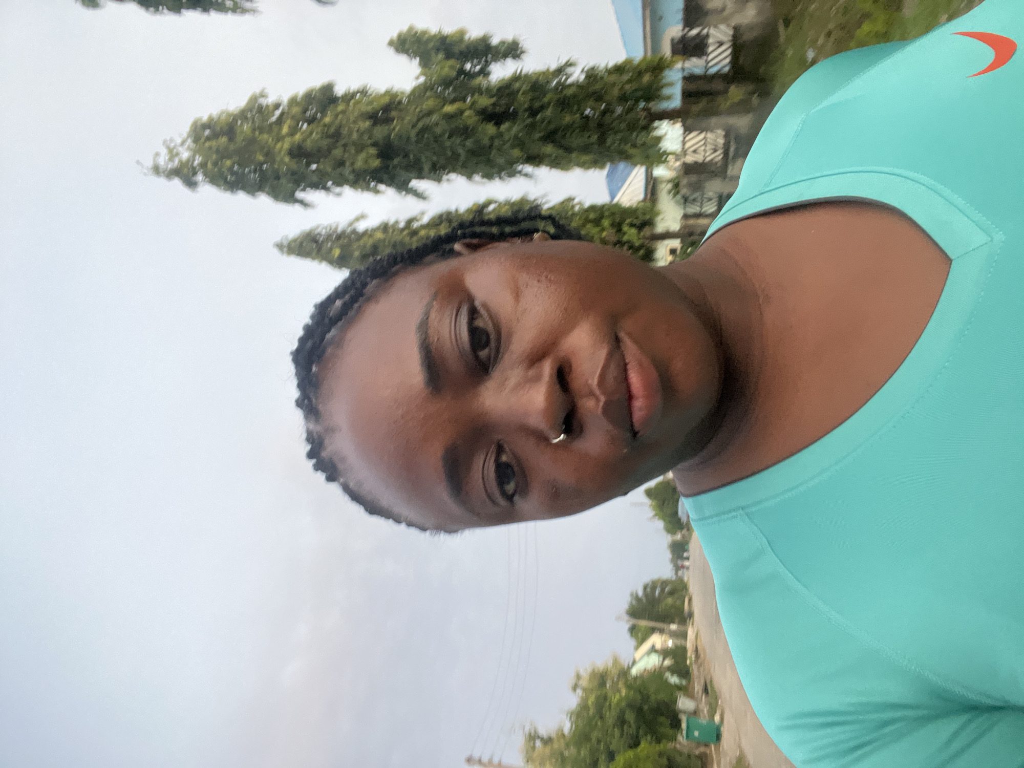 A medium shot photograph shows a Black woman from the chest up, facing the camera directly. She has dark braided hair, brown eyes, and a silver nose ring. She is wearing a teal V-neck shirt with a small orange swoosh logo on the right side of her chest. Her expression is neutral. The background is outdoors, with a street and some trees visible. The sky is overcast. The image appears to have been taken in a residential area in Abuja, Nigeria, given the style of the houses and the vegetation.