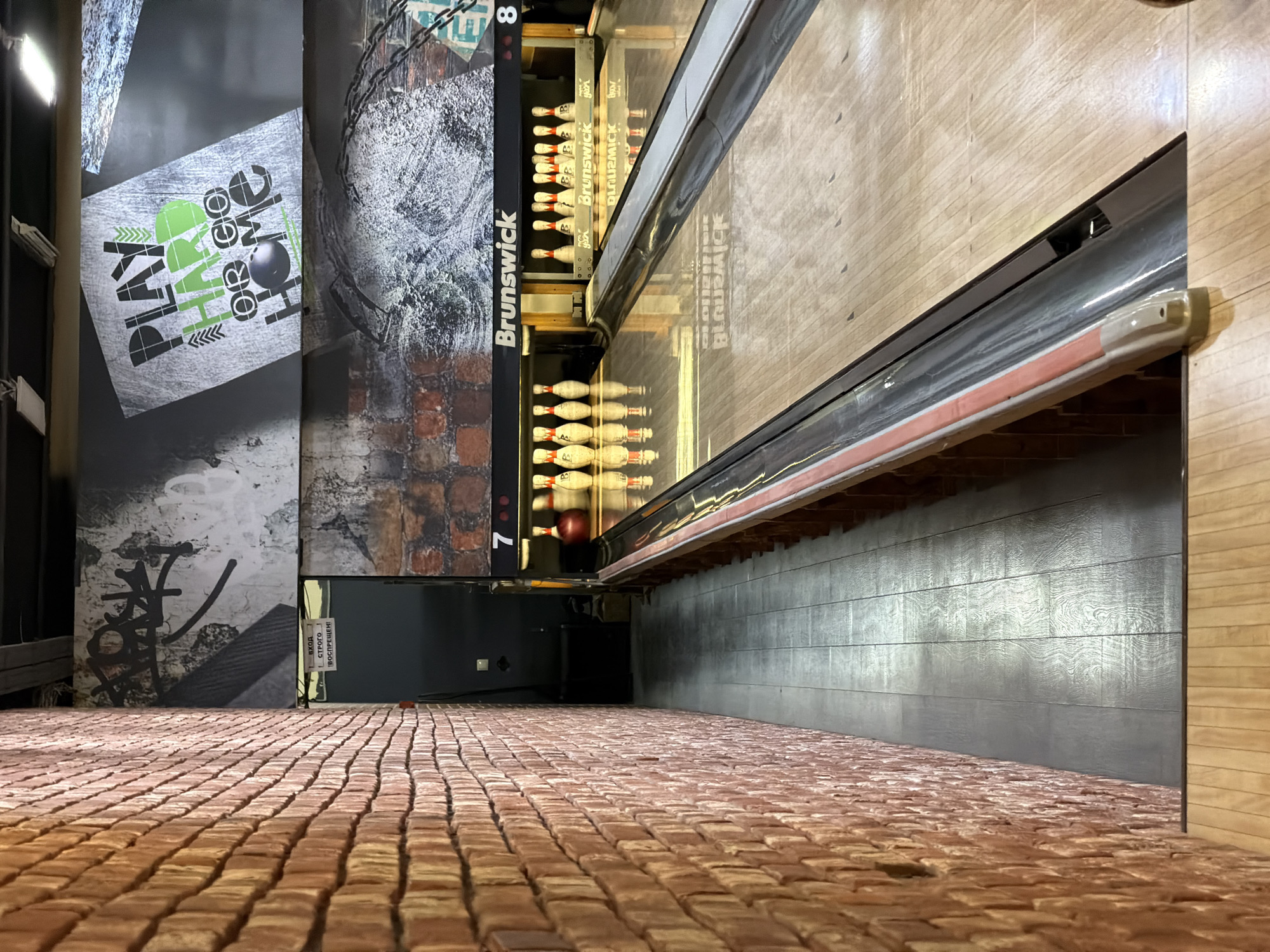 The image shows a bowling alley in Moscow, Russia. A brick wall is on the left side of the frame, and a bowling lane stretches into the distance on the right. There are bowling pins set up on the lane, and a red bowling ball is on the approach. There are posters on the wall behind the bowling lane. One poster has the text "PLAY HARD OR GO HOME" in large letters. Another poster has a picture of graffiti. A sign above a doorway reads "ВХОД СТРОГО ВОСПРЕЩЁН!" which translates to "ENTRANCE STRICTLY PROHIBITED!" The bowling lane is marked with "Brunswick" on the side.