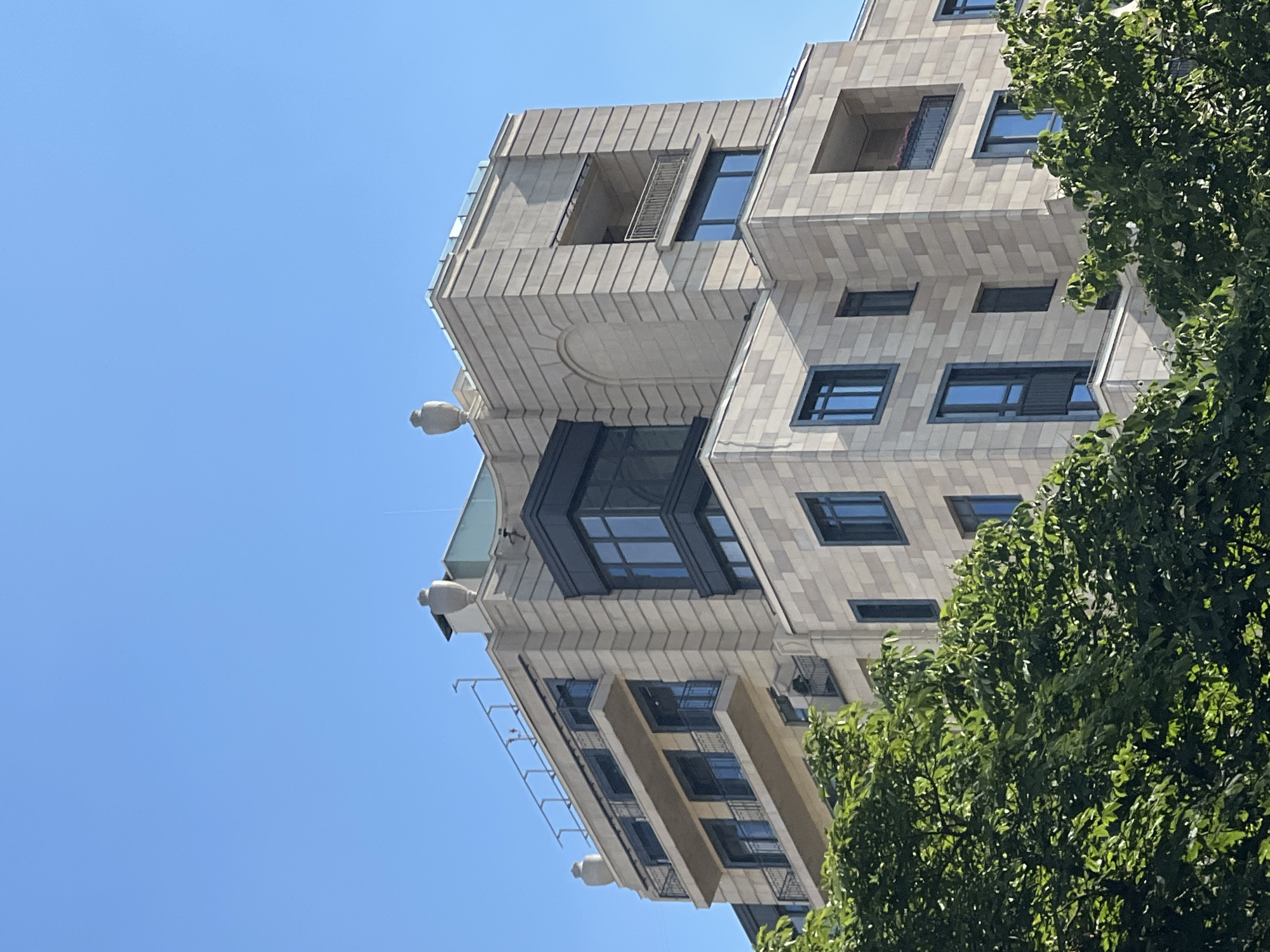 This low-angle, vertically oriented image shows a modern apartment building with a light-colored stone facade against a clear blue sky. The building features a geometric design with a mix of rectangular windows, some with dark frames and others recessed within the facade. Balconies with dark railings are visible on lower floors, and the upper sections of the building have protruding elements and open-air spaces. Decorative gargoyle-like figures are mounted on the building's upper levels.
To the right, a lush green tree with dense foliage partially obscures the building. The sunlight suggests it is daytime, likely late morning or early afternoon, given the bright, shadowless illumination. The clear sky indicates fair weather. The overall scene conveys a sense of contemporary urban architecture and a pleasant, sunny day. No people are visible in the image, and there is no visible text. The context provided indicates the location is Moscow, Russia, which aligns with the architectural style often seen in upscale urban developments.