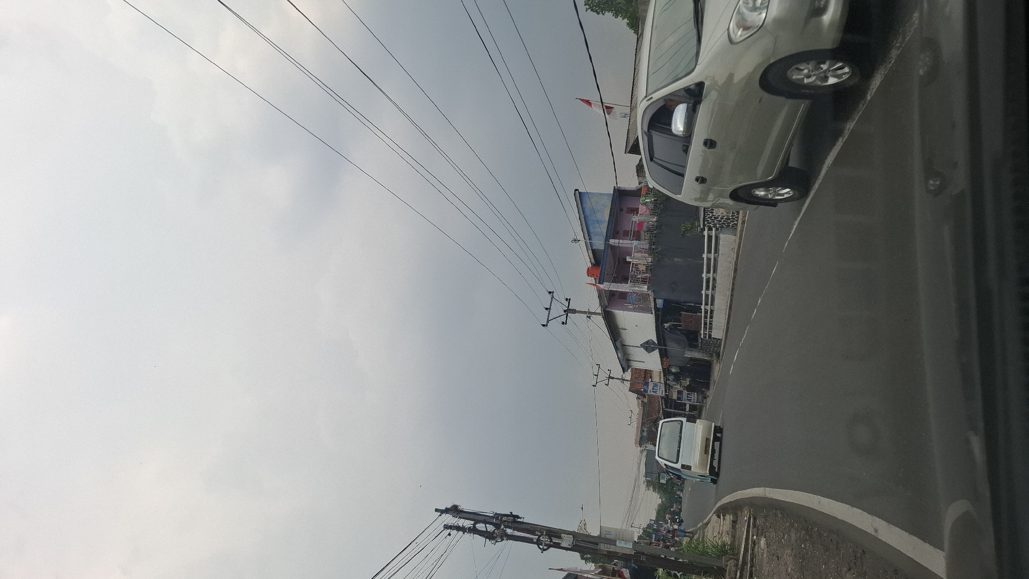 This is a vertical, eye-level, outdoor, daytime, full-shot photograph taken from a vehicle's perspective, looking forward along a paved road in Sukasari, Indonesia. The sky is overcast with light gray clouds, indicating a cloudy or possibly rainy day. The road curves slightly to the left and ahead.
On the left side of the road, a narrow concrete sidewalk runs along the edge, bordered by dry grass and a few small plants. A utility pole with numerous wires is prominently featured on the left, leaning slightly to the right. Behind the sidewalk, there are several buildings and a few people walking. One building appears to have "ATM" signage. Further down the road, more buildings and vehicles are visible.
On the right side of the road, a silver minivan is in motion, facing away from the camera and slightly to the left. The minivan's headlights are on. Further ahead, another building with a multi-colored facade (pink, blue, and white) is visible. Indonesian flags are displayed on poles attached to this building. A white and red flag is clearly visible. There are other structures and overhead wires throughout the scene. The overall impression is of a residential or commercial street in a built-up area under a cloudy sky.