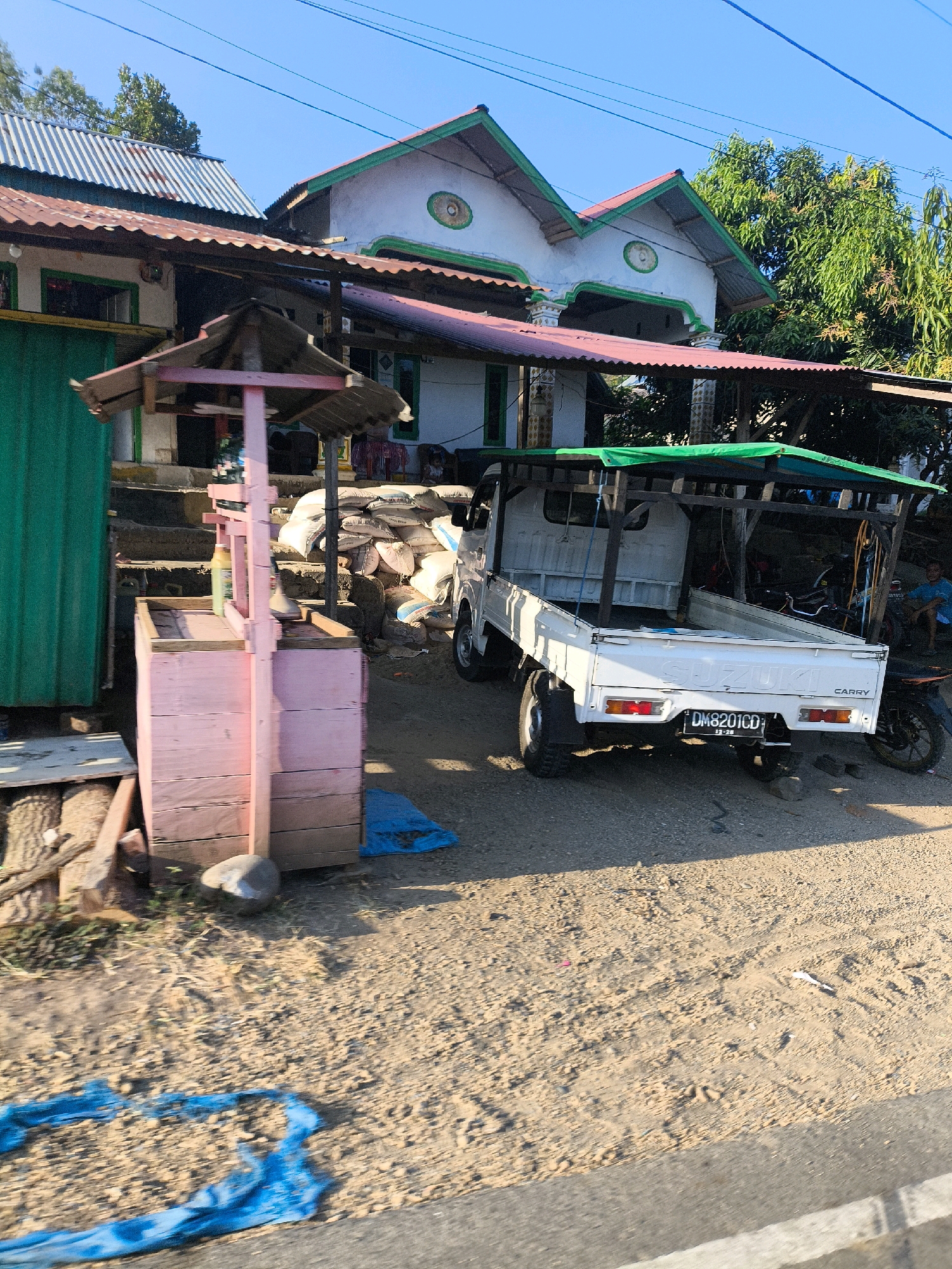 The image depicts a street scene in Pentadu Timur, Indonesia, under a clear blue sky. In the foreground, a dirt and gravel road is visible, with a blue tarp spread out and some scattered debris. To the left, a light pink wooden stand with a grey corrugated roof is positioned near a green corrugated metal wall. Behind the stand, a set of concrete stairs leads up to a building with a red corrugated metal roof.
Further back, a white Suzuki Carry truck is parked on the right side of the road. The truck has a green tarp covering its bed. Stacked white sacks are visible behind the truck. A motorcycle is parked to the right of the truck, and a man in a blue shirt is sitting near it. The buildings in the background have white walls and red corrugated metal roofs with green trim. Trees with dense foliage are visible behind the buildings. The overall impression is of a sunlit, possibly rural or semi-urban environment.