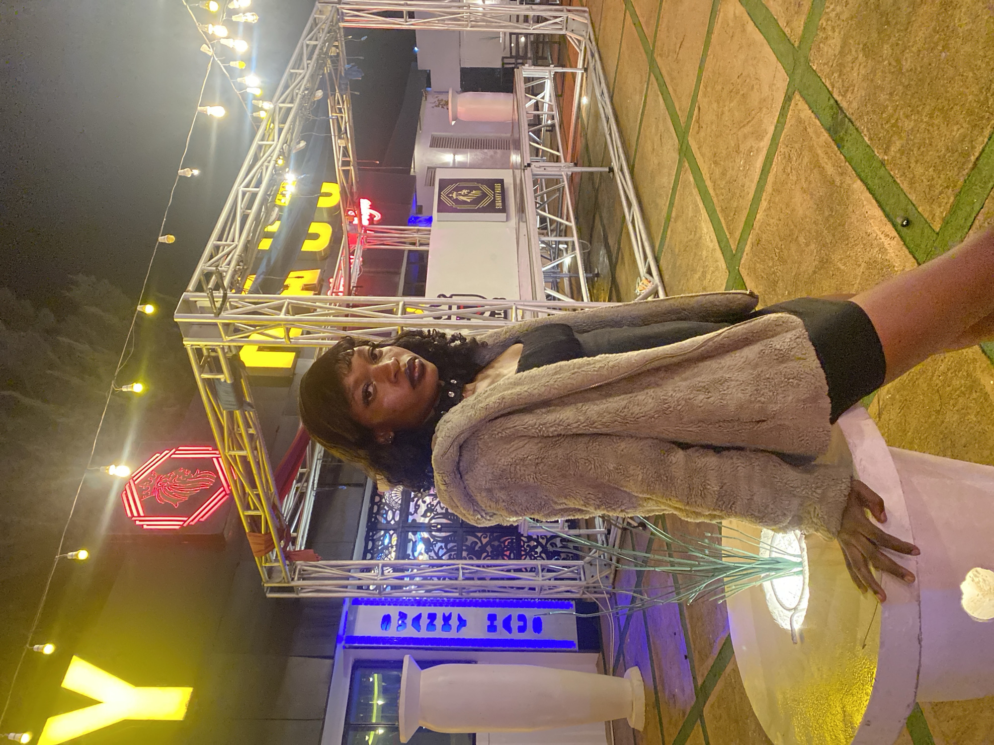 The image depicts an outdoor nighttime scene, possibly in the city of Gura Top, Nigeria. A young Black woman is positioned in the foreground, leaning on a white circular planter with a plant inside. She is wearing a fuzzy tan hooded jacket over a black dress and a black choker necklace. Her gaze is directed towards the camera. Behind her, a metallic stage structure is visible, adorned with yellow and red neon signs. One sign spells out "SWANKY HAUS" in white text with a lion logo. String lights hang above the structure, illuminating the area. The ground is paved with large, square tiles in a yellowish-brown color, with green lines separating them. To the left, a tall, white vase-like structure is illuminated by blue light. The overall ambiance suggests a lively entertainment venue.
